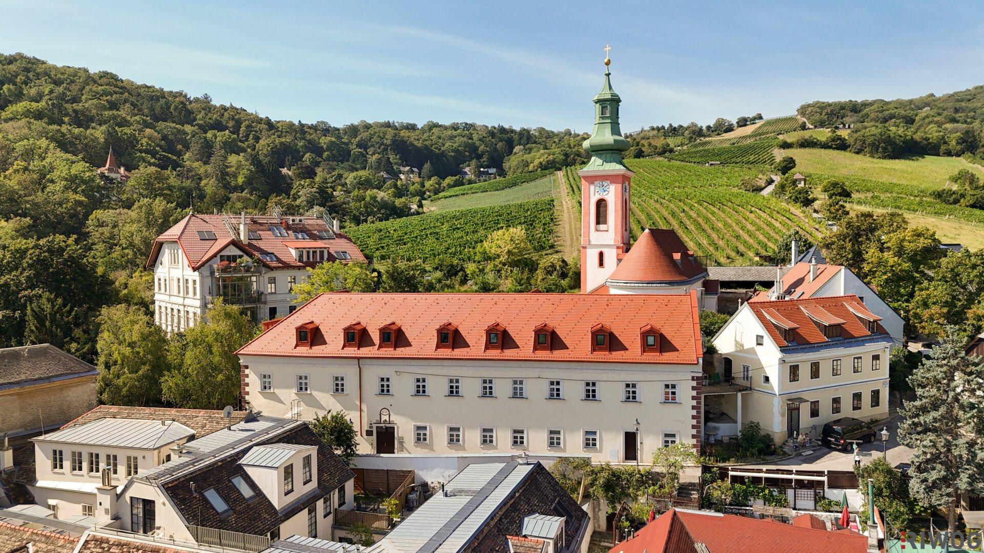 Historisches Gebäudeensemble mit Kirche und Weinbergen in einer malerischen, grünen Landschaft.