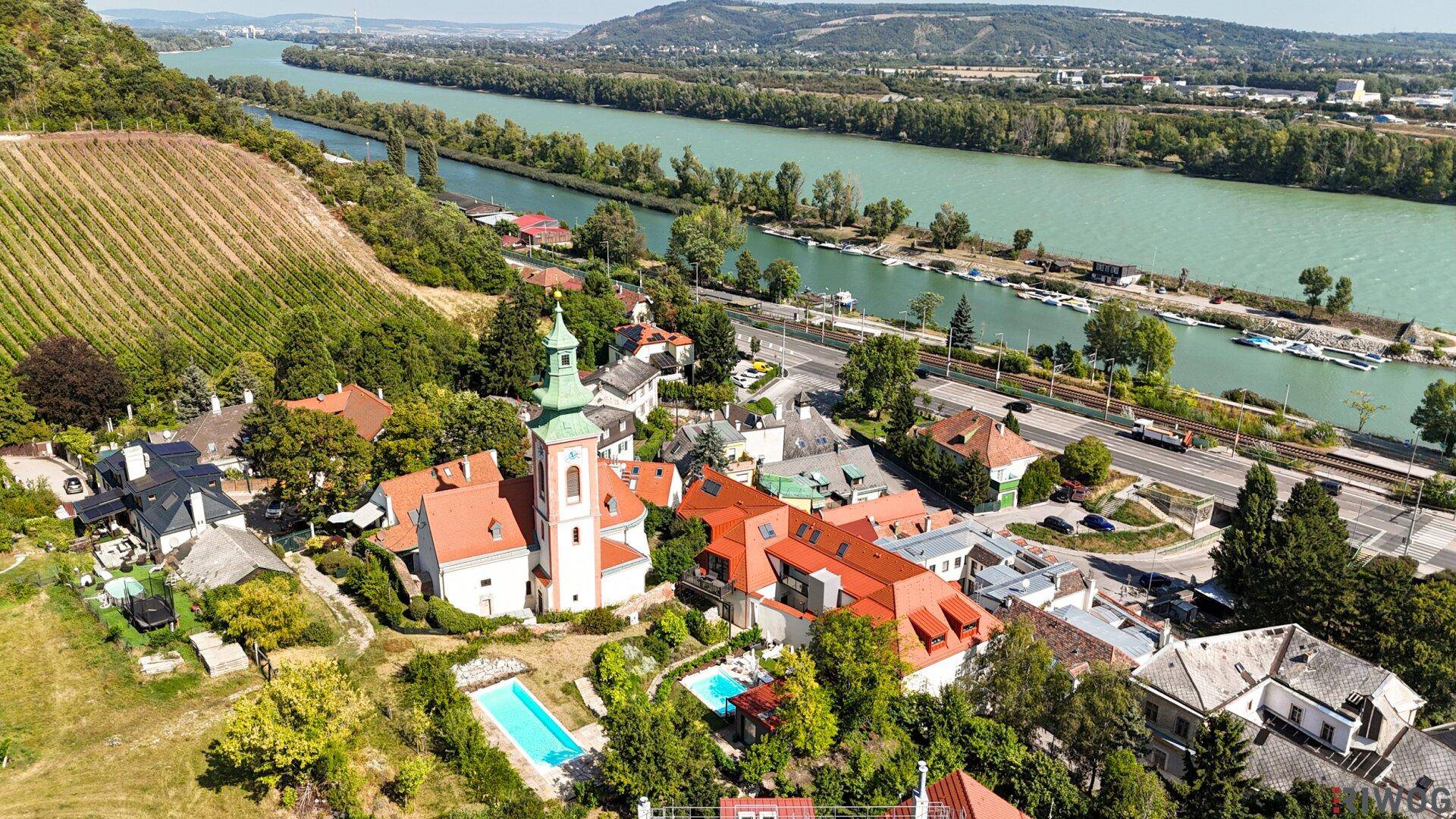 Luftaufnahme eines Dorfes am Fluss mit Kirche, Weinbergen, Pools und umliegender Natur.