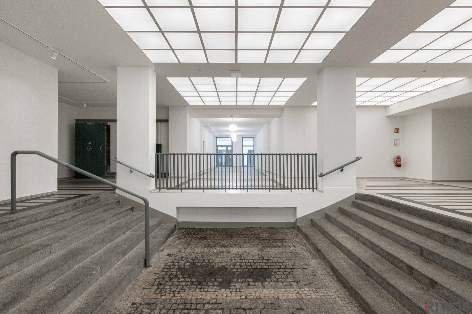 Heller Eingangsbereich mit breiten Treppen, Steinboden und einer modernen Deckenbeleuchtung.