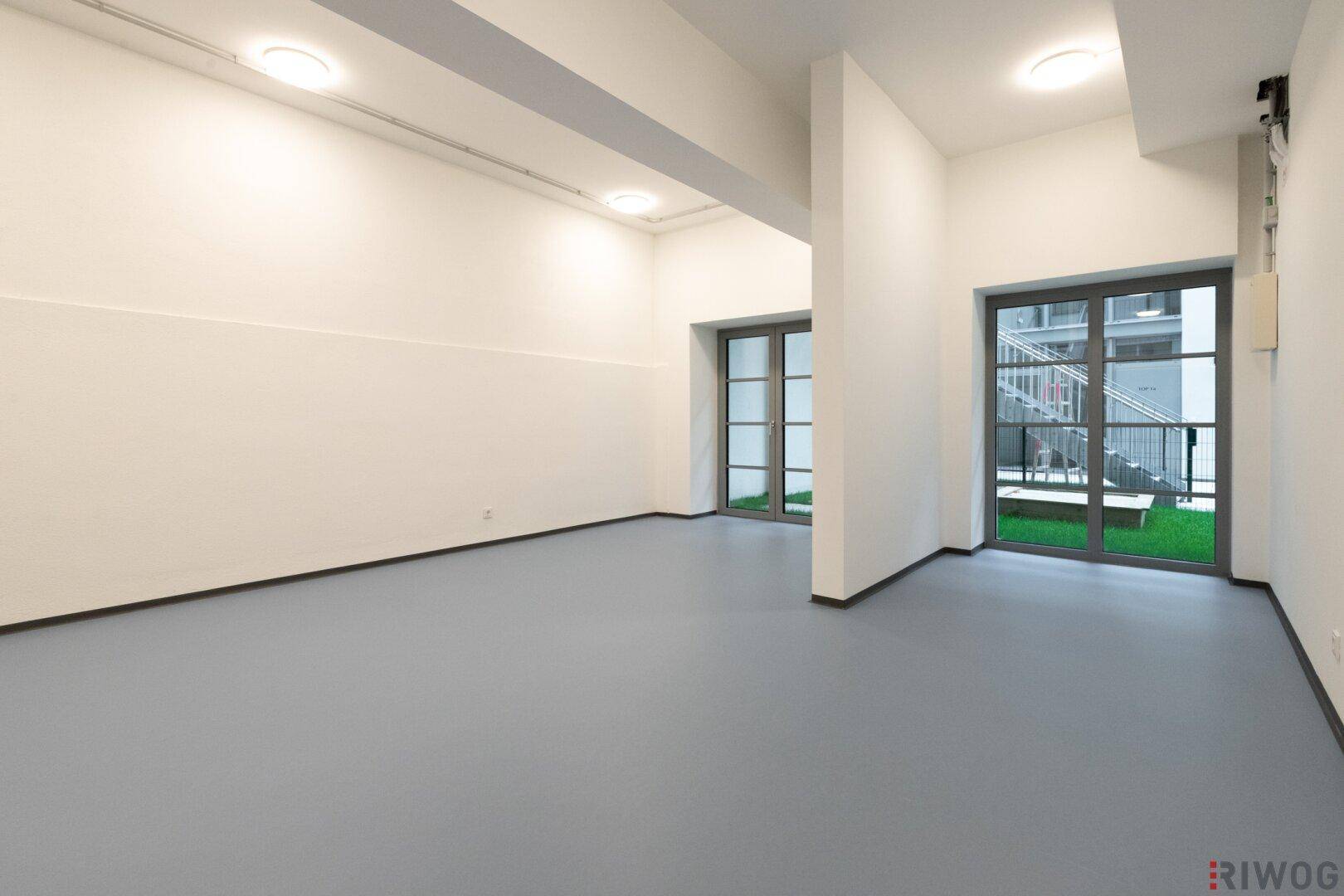Heller, leerer Raum mit großen Fenstern und Zugang zu einem kleinen Außenbereich.