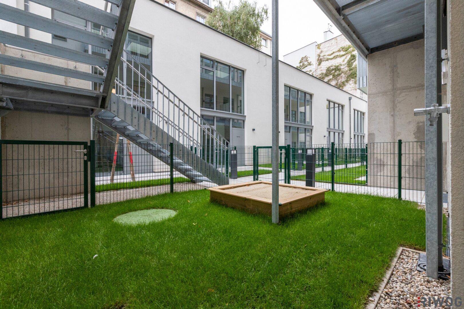 Gemeinschaftsgarten mit Rasenfläche, Zaun und einem Sandkasten für Kinder.