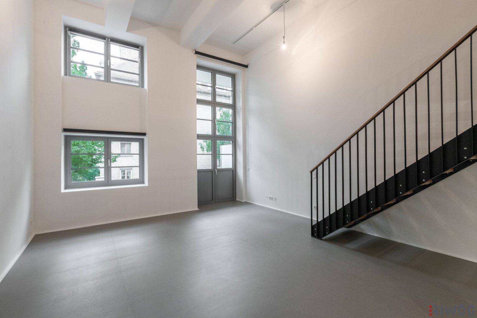 Heller Wohnraum mit hohen Decken, großen Fenstern und einer eleganten Treppe.