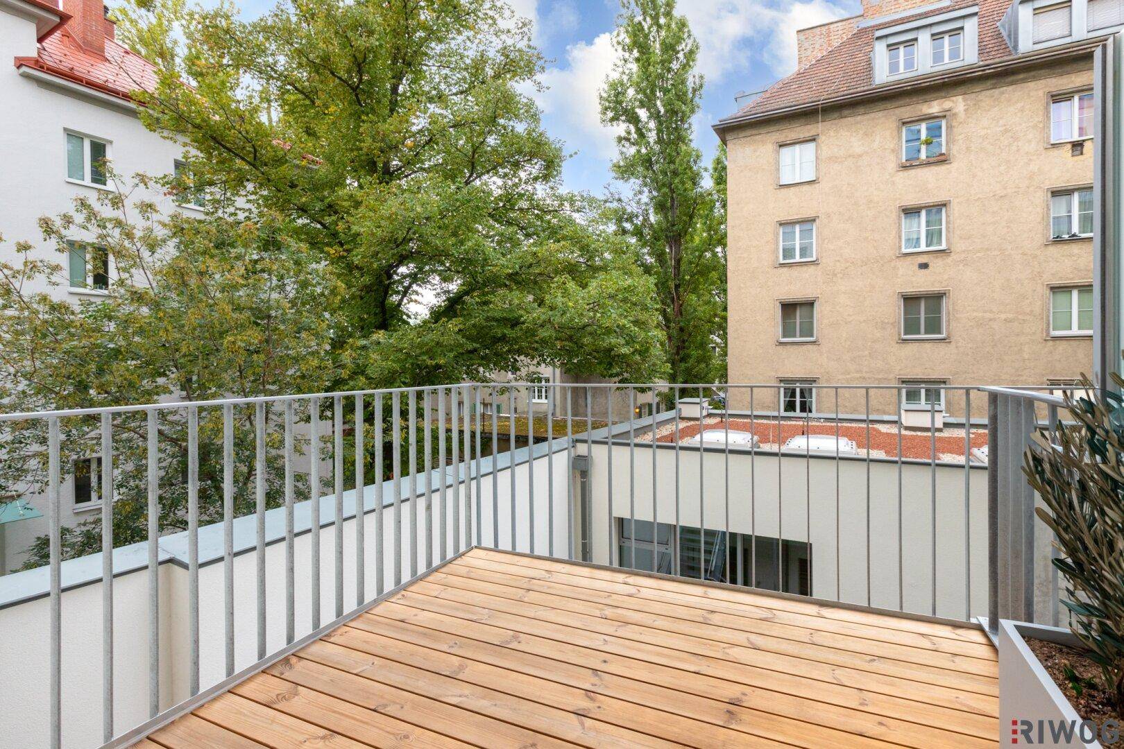 Heller Balkon mit Holzfußboden und Metallgeländer, umgeben von Bäumen und Gebäuden.