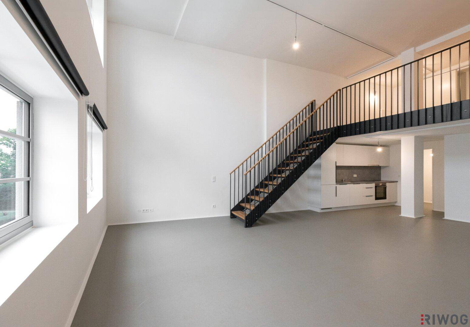Geräumiger, unmöblierter Loft-Wohnbereich mit hohen Decken, großen Fenstern und Blick auf die Galerie.