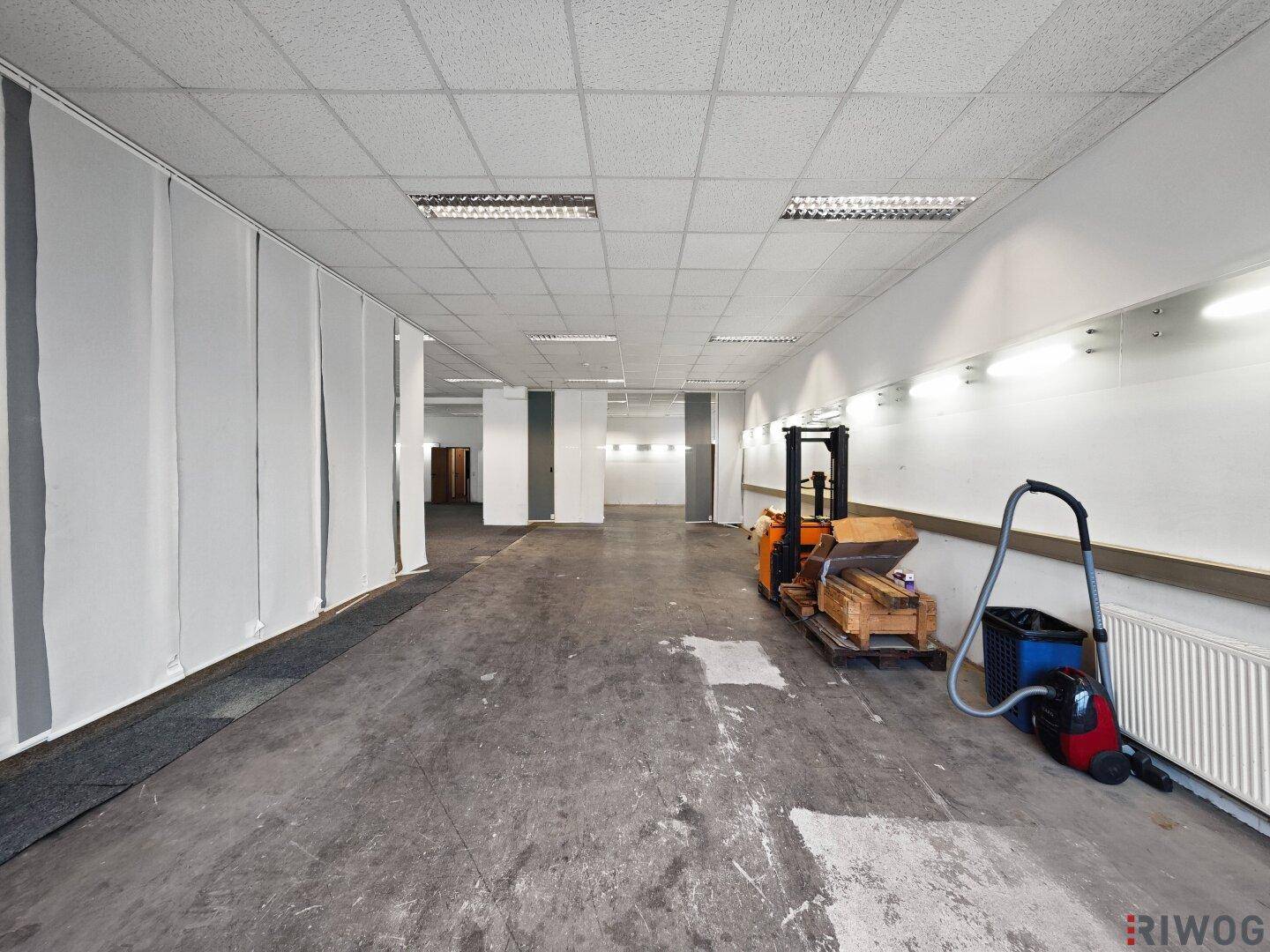 Geräumiger Gewerberaum mit Betonboden und Teppichstreifen, ausgestattet mit Heizkörpern und Beleuchtung.
