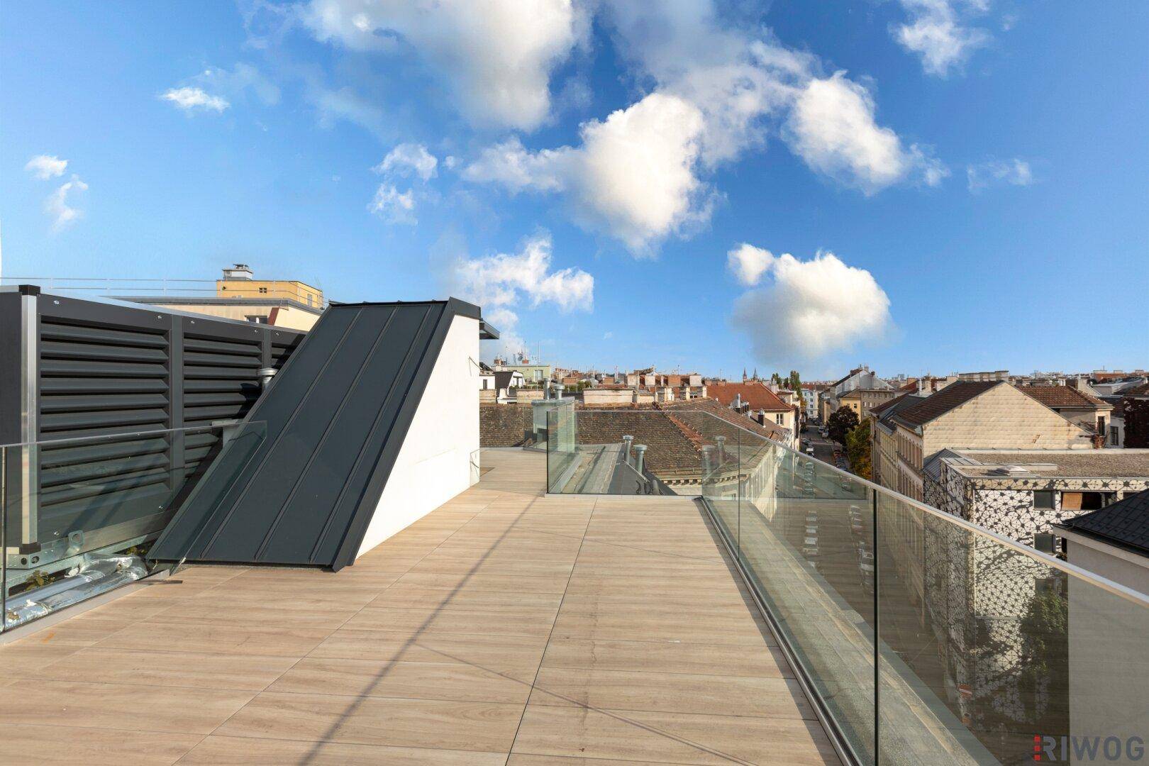 Weitläufige Dachterrasse mit Holzfliesen und Glasgeländer, umgeben von Stadtblick und blauem Himmel.