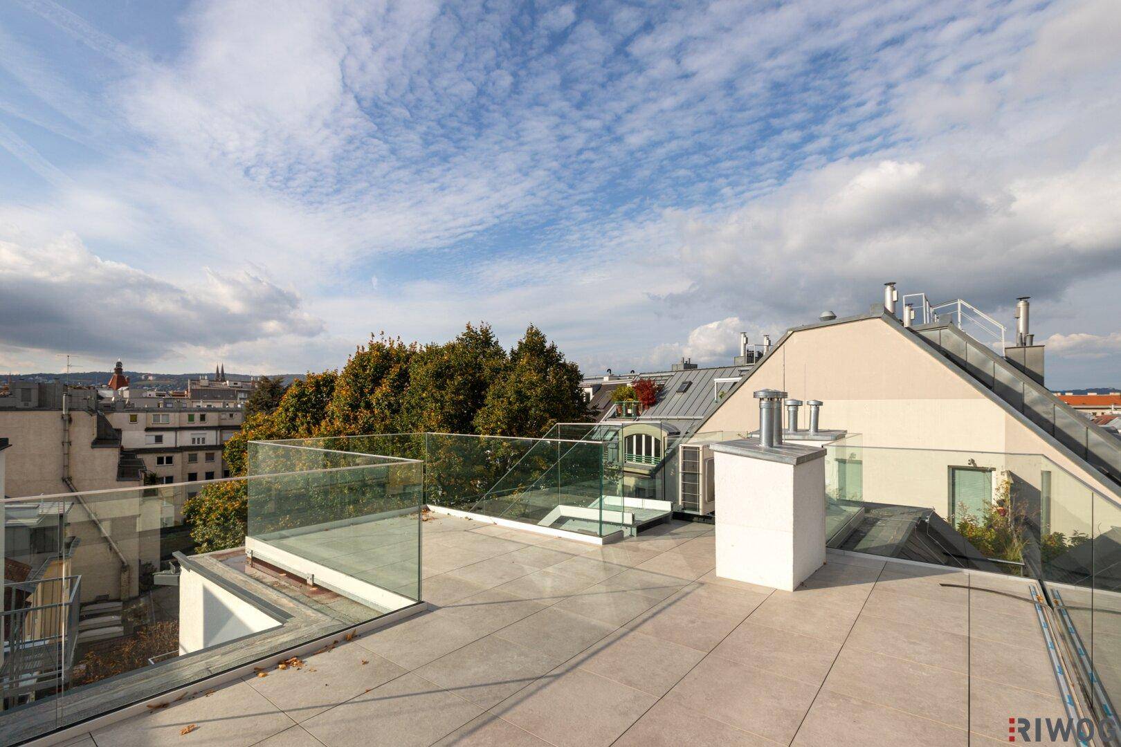 Eine weitläufige, unmöblierte Dachterrasse mit Glasgeländer und Blick über die Stadt.