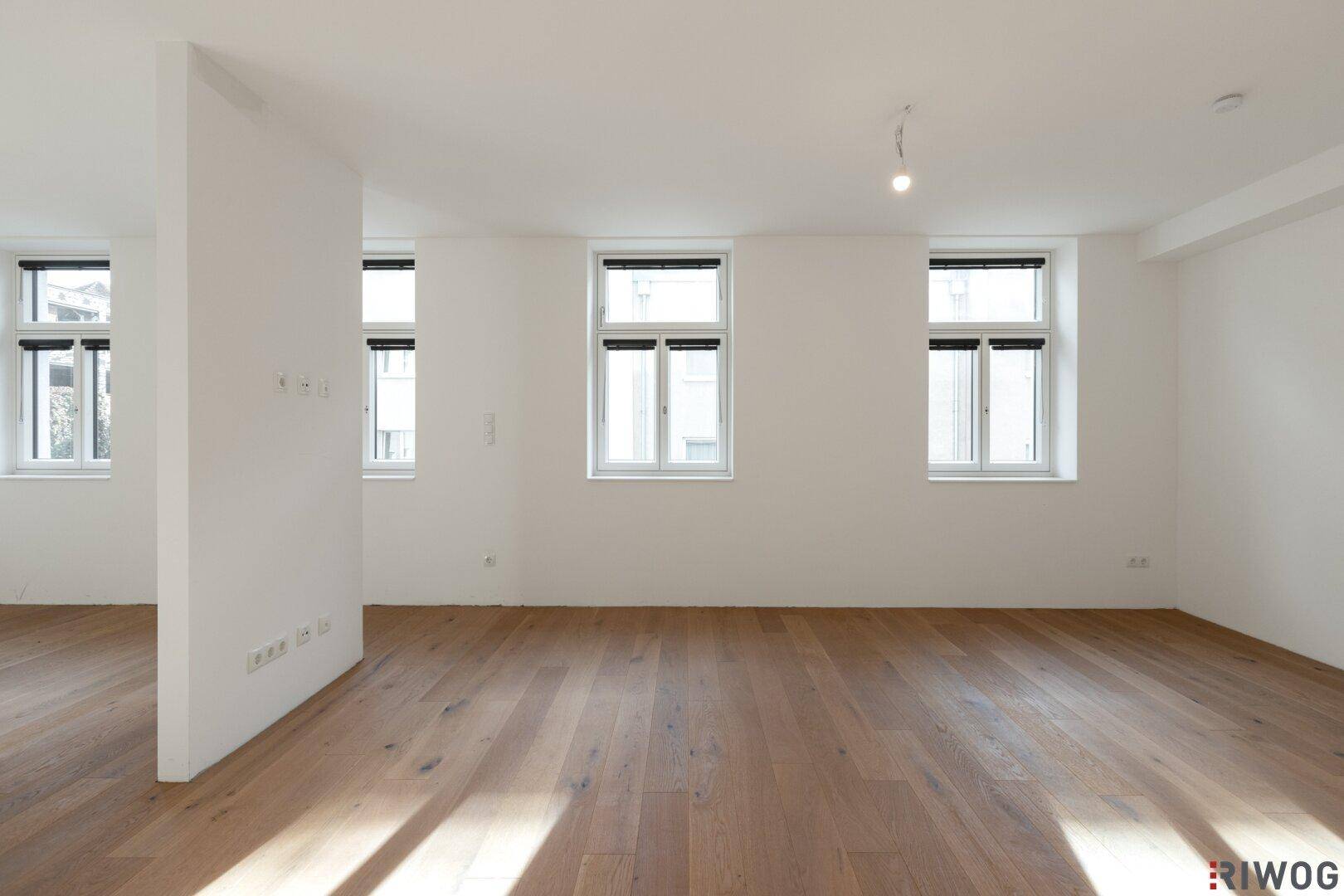 Geräumiger, heller Wohnbereich mit großen Fenstern und elegantem Holzboden, bereit zur individuellen Gestaltung.