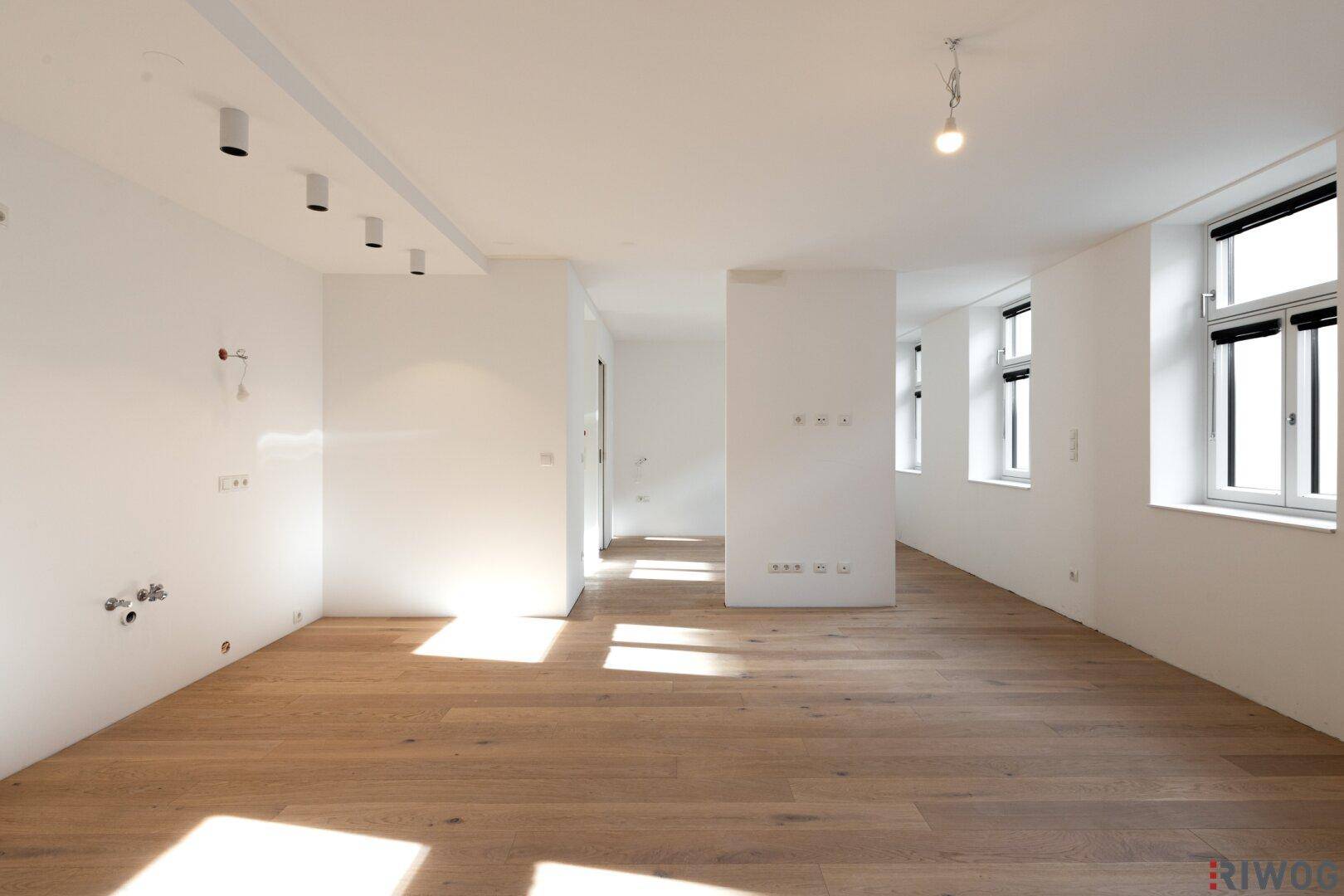Geräumiger, heller Raum mit Holzboden und vorbereiteten Anschlüssen für eine Küche oder einen Hauswirtschaftsraum.