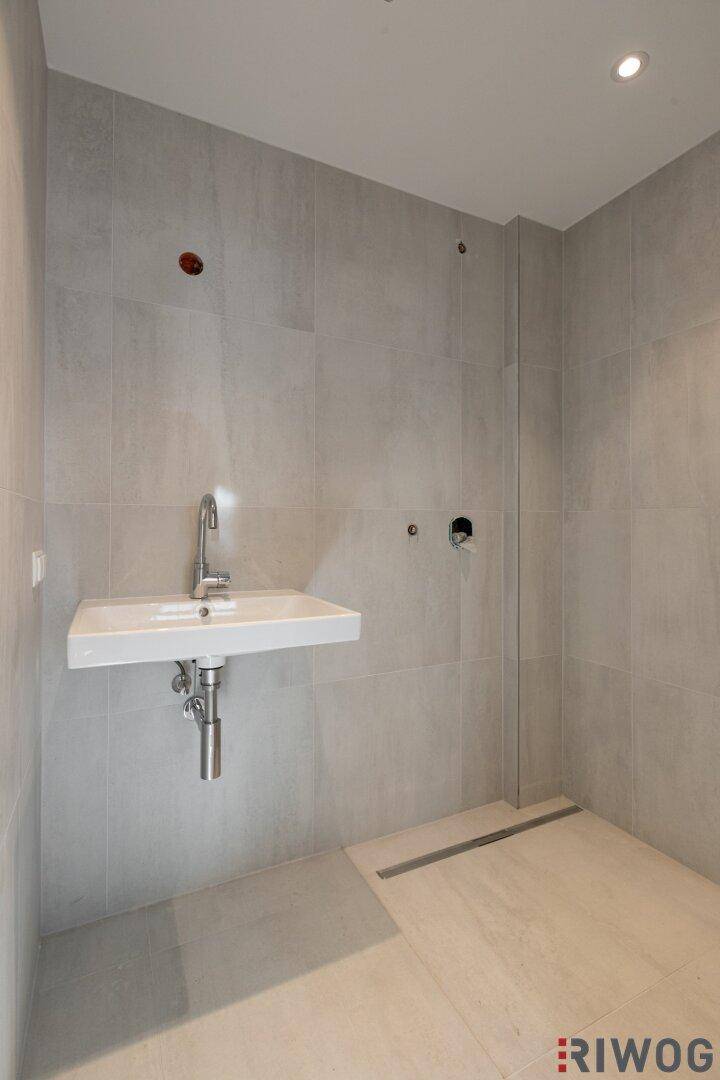 Modernes Badezimmer mit grauen Fliesen, wandmontiertem Waschbecken und vorbereiteter begehbarer Dusche.