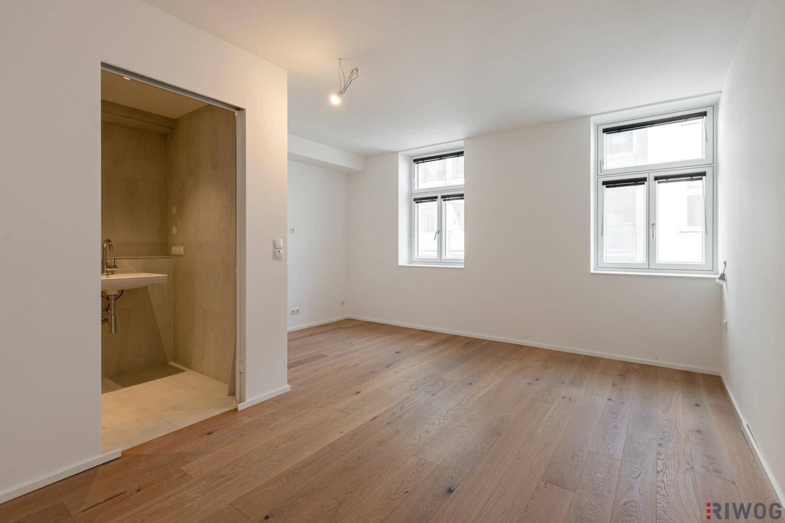 Leeres Schlafzimmer mit Holzboden, großen Fenstern und offenem Waschbereich, bereit zur Einrichtung.