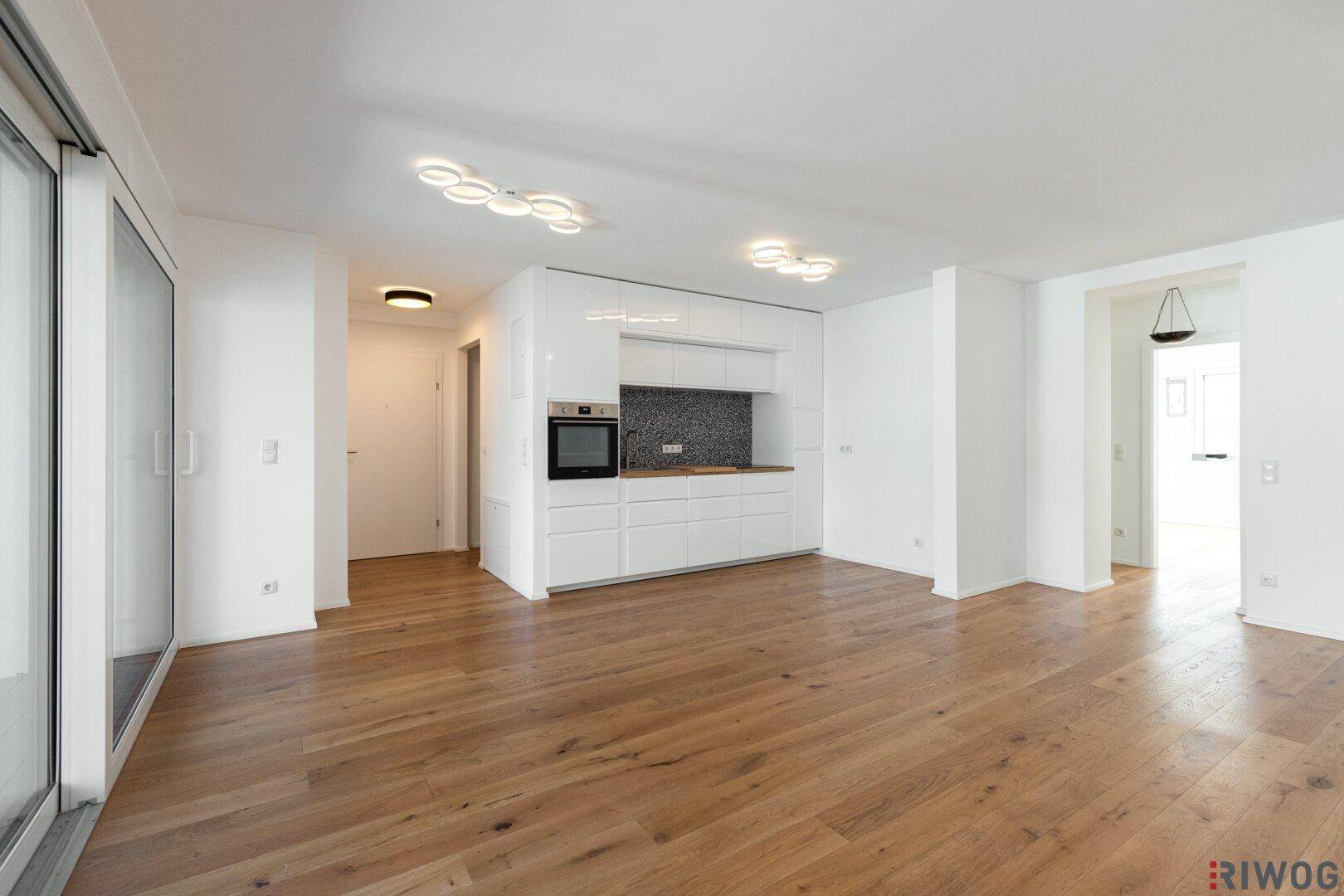 Geräumiger, leerer Wohnbereich mit offener Küche, Holzboden und viel Tageslicht.