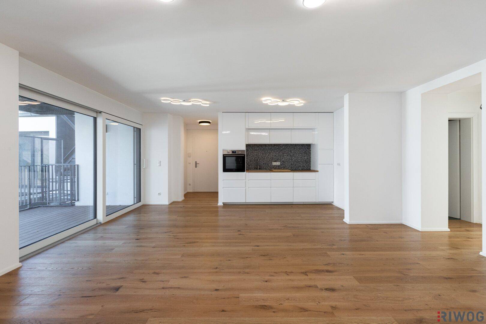 Weitläufiger, leerer Wohnbereich mit integrierter Küche, Holzboden und Schiebetüren zur Terrasse.