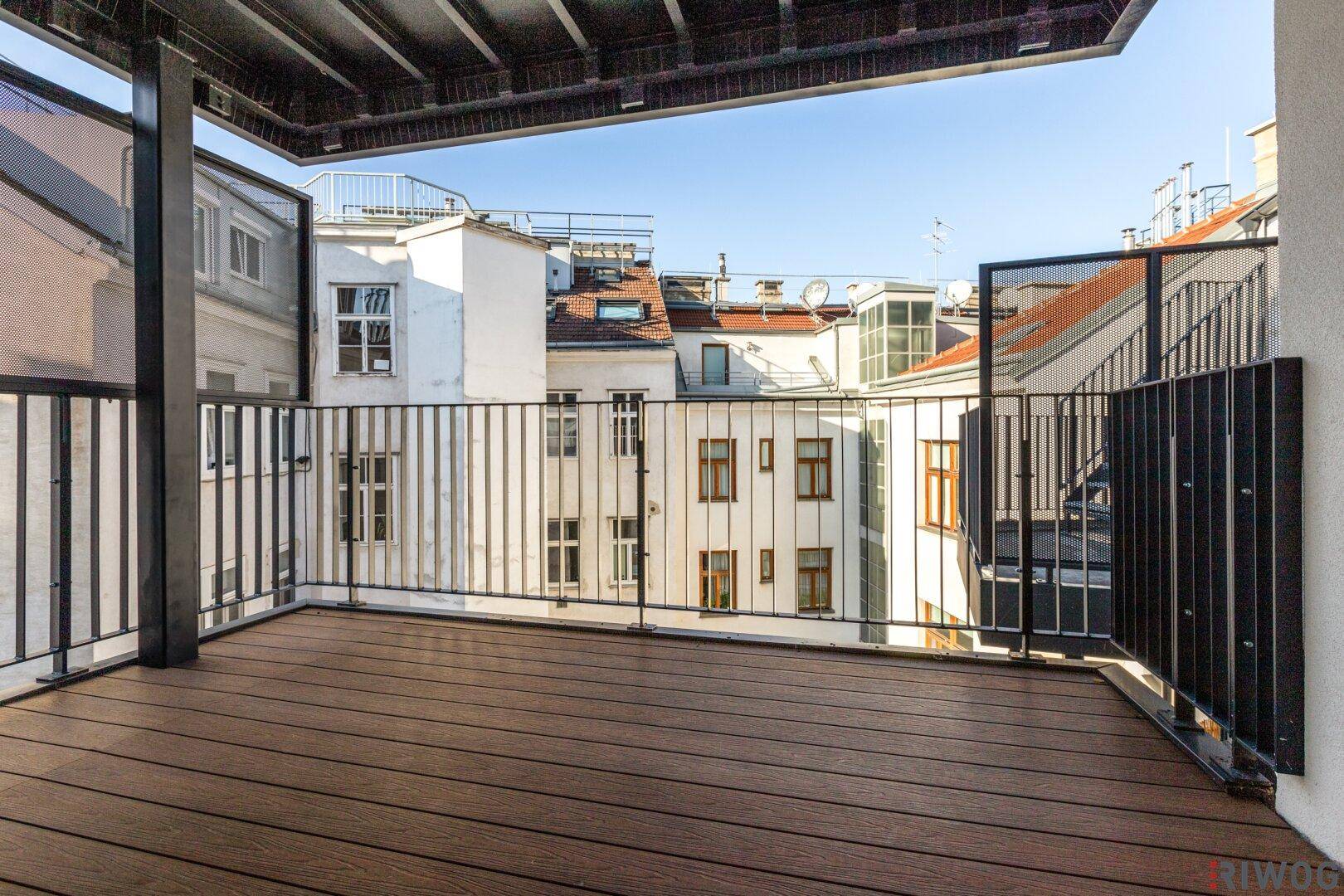 Große, leere Terrasse mit Holzboden und Metallgeländer, umgeben von Stadtgebäuden.