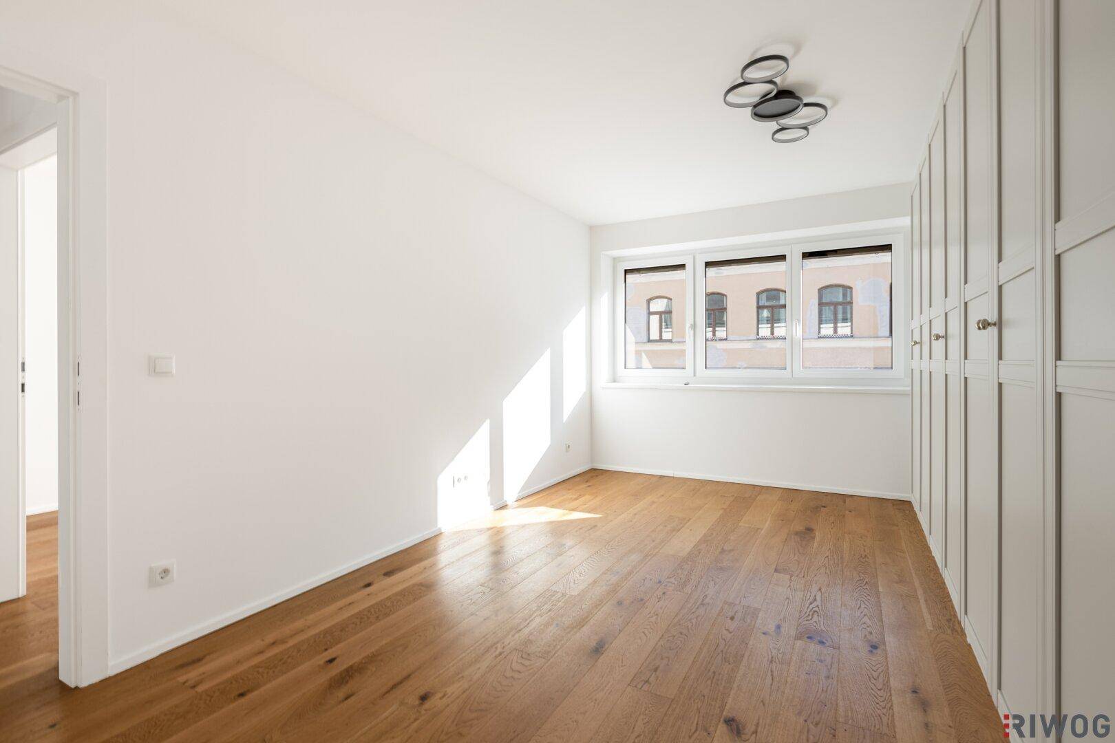 Leeres Schlafzimmer mit Holzboden, weißen Wänden und großem Fenster.