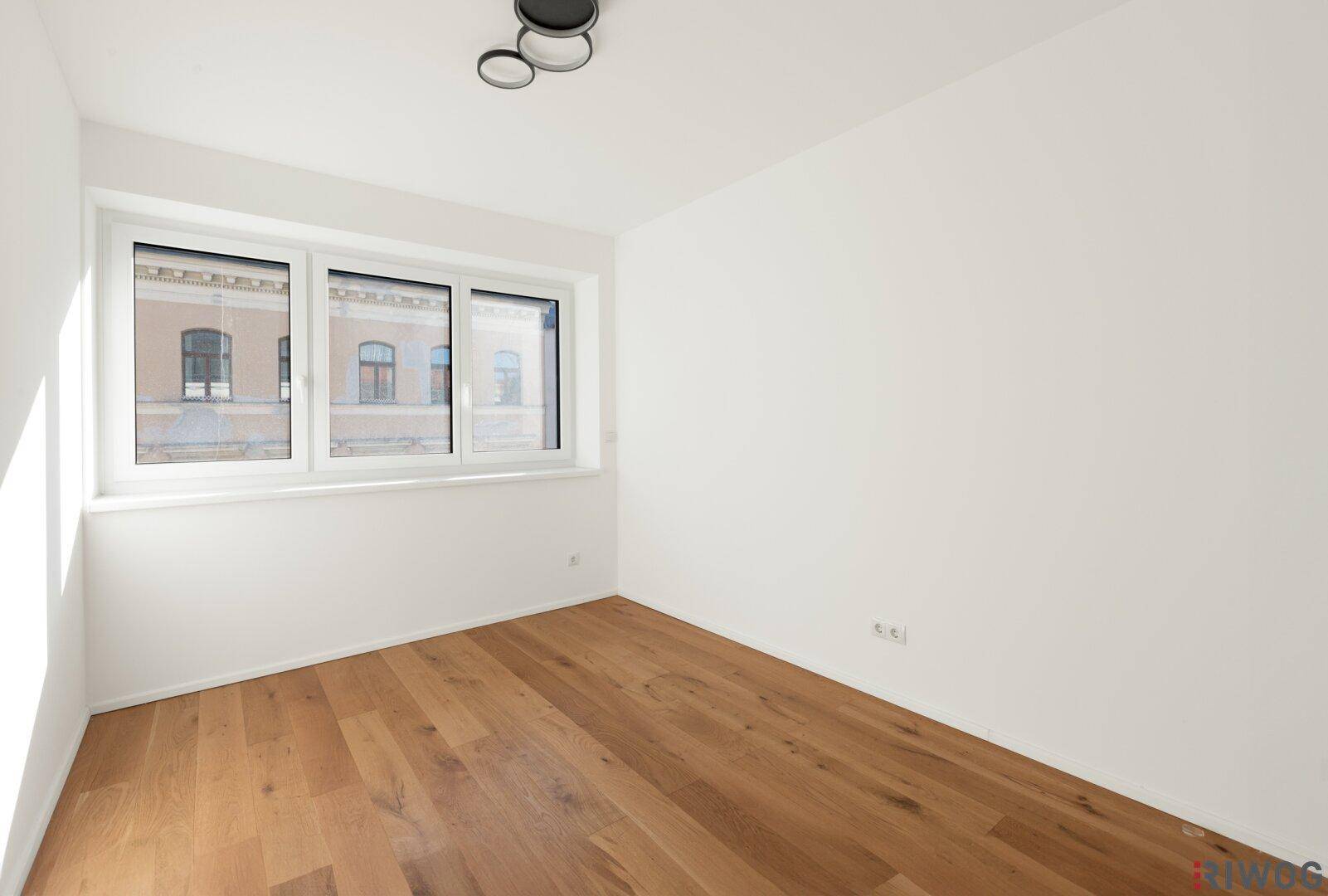 Kleineres Zimmer mit Holzboden, weißen Wänden und Fenster zur Straße.