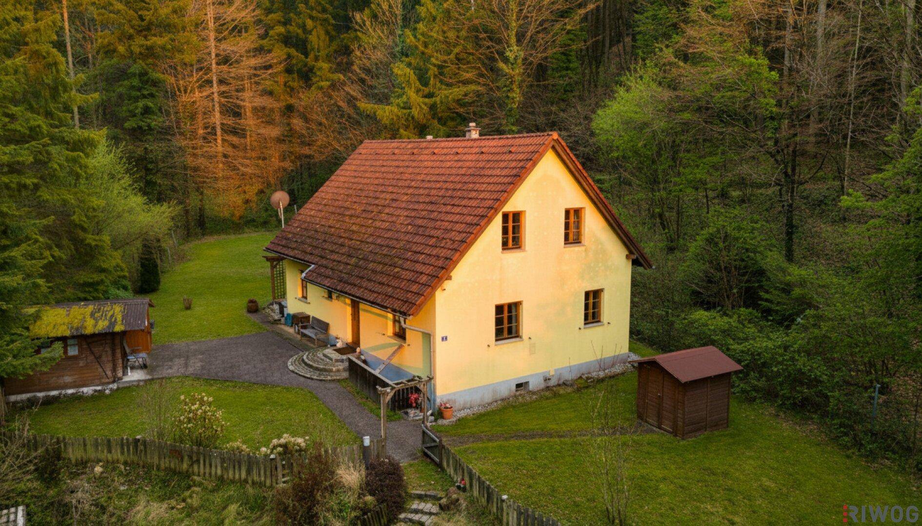 Nahaufnahme des gelben Hauses mit rotem Satteldach, umgeben von einem gepflegten Garten und Bäumen.