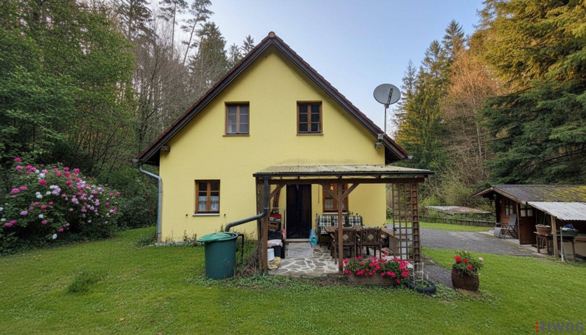 Die Hausfassade mit überdachter Terrasse und Gartenmöbeln, umgeben von grüner Natur.