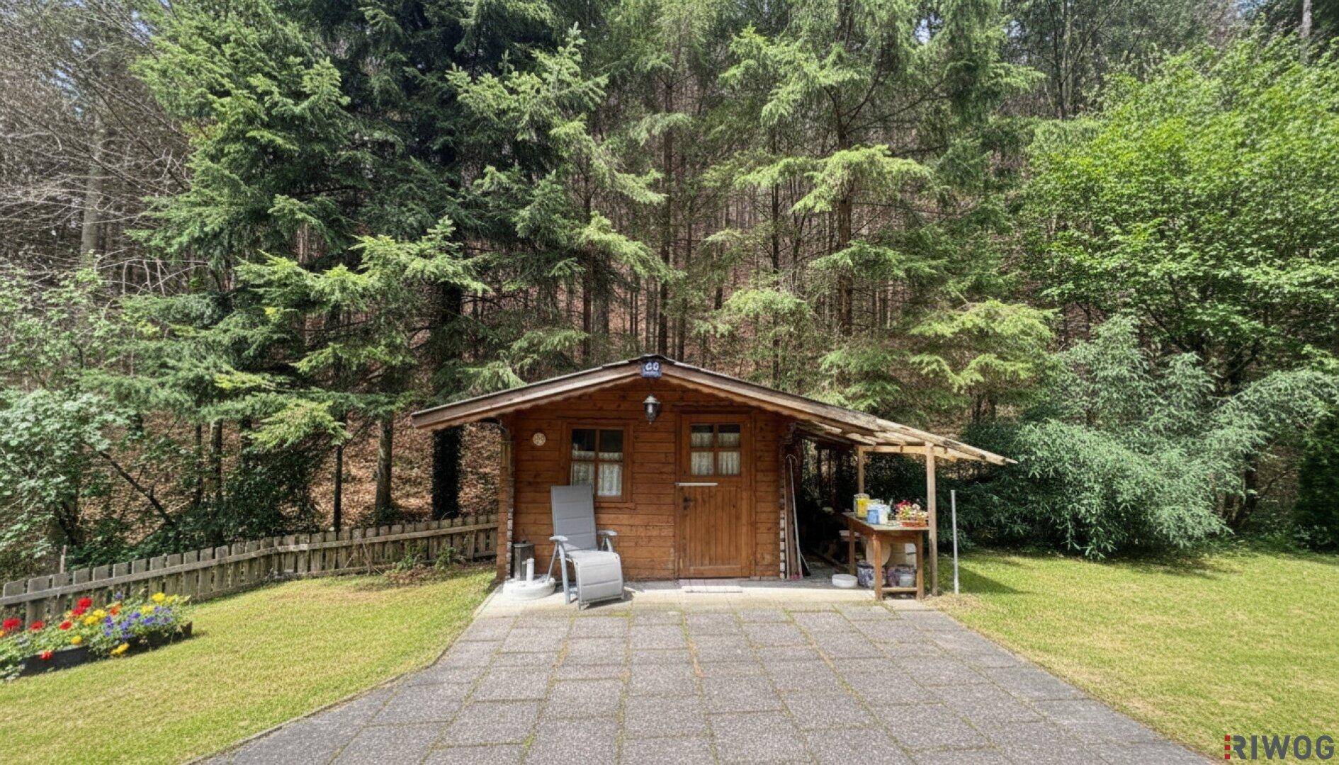Ein rustikaler Holzschuppen mit gepflastertem Vorplatz, umgeben von Rasen und Wald.