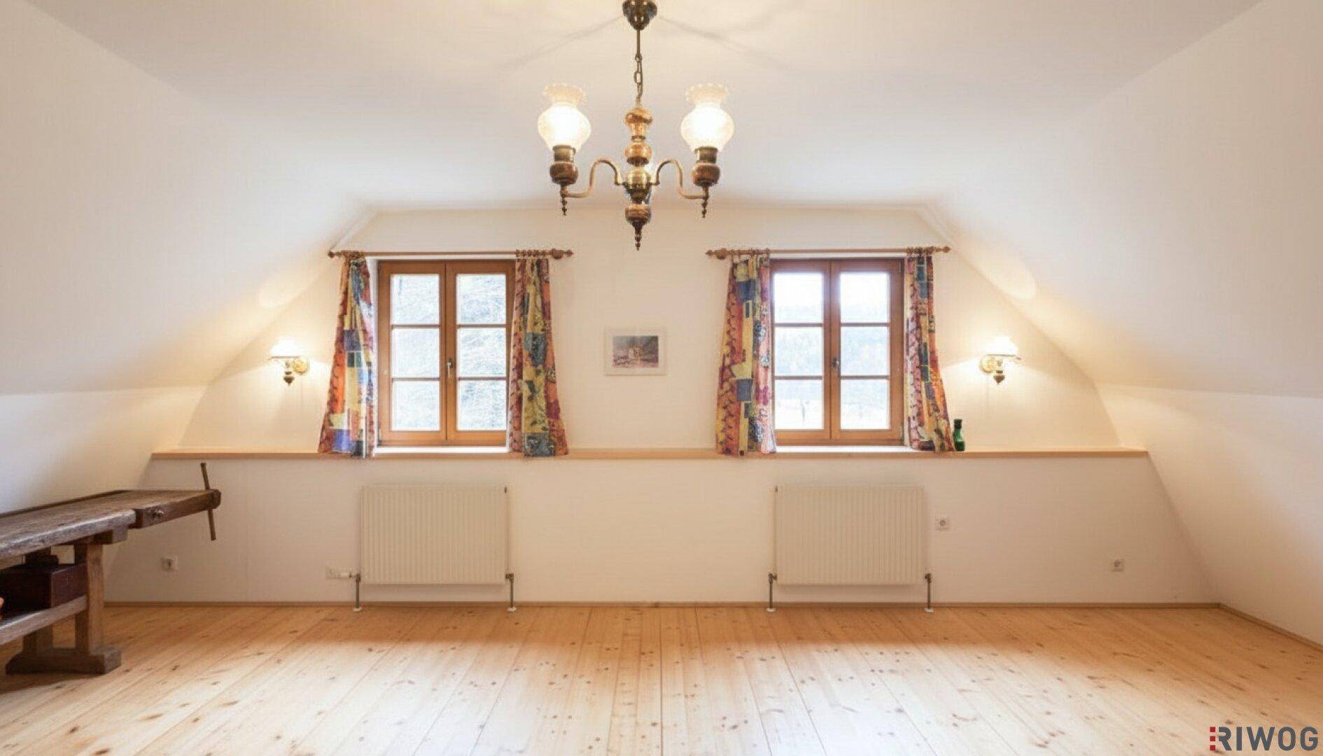 Heller Dachgeschossraum mit Holzboden, Dachschrägen und zwei Fenstern, die viel Licht hereinlassen.