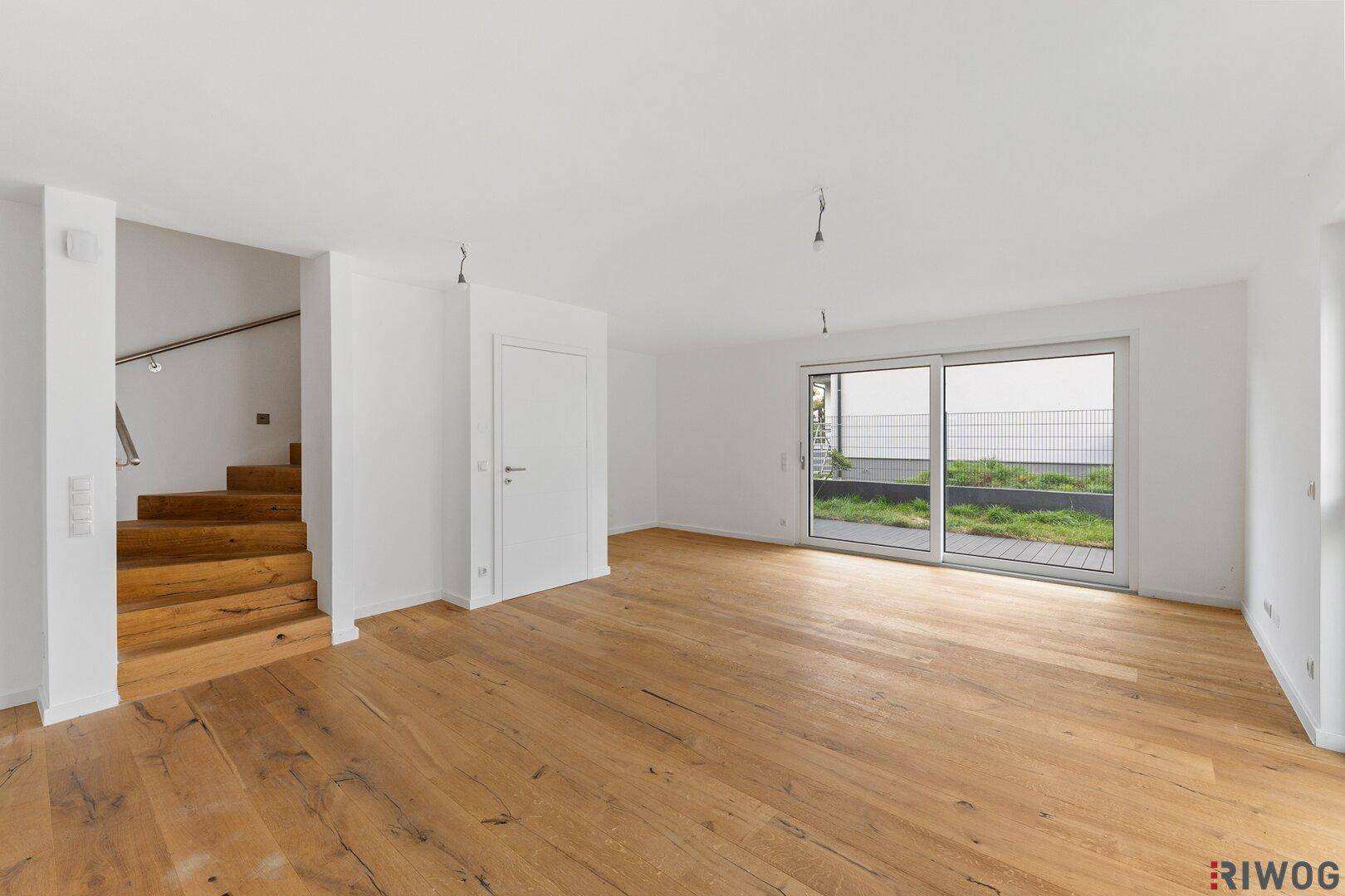 Helles, leeres Wohnzimmer mit Holzboden, großen Fenstern und Zugang zur Terrasse.