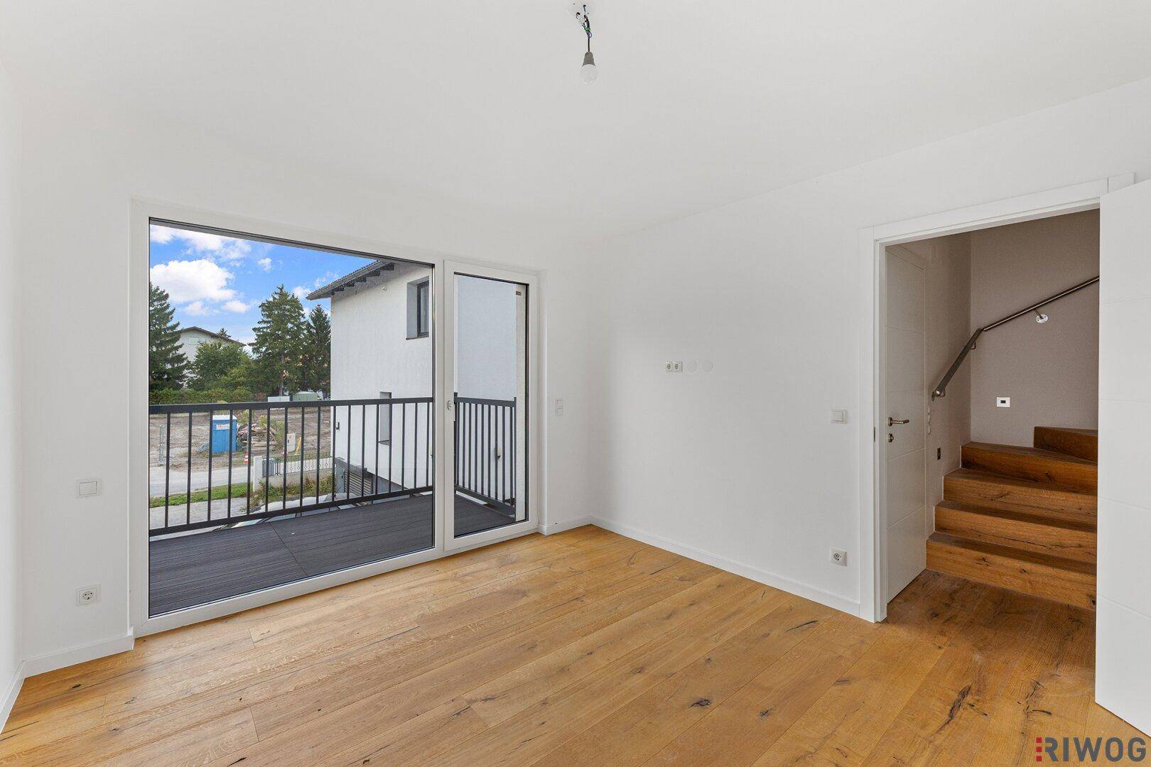 Helles, leeres Zimmer mit Holzboden und großem Fenster zum Balkon.