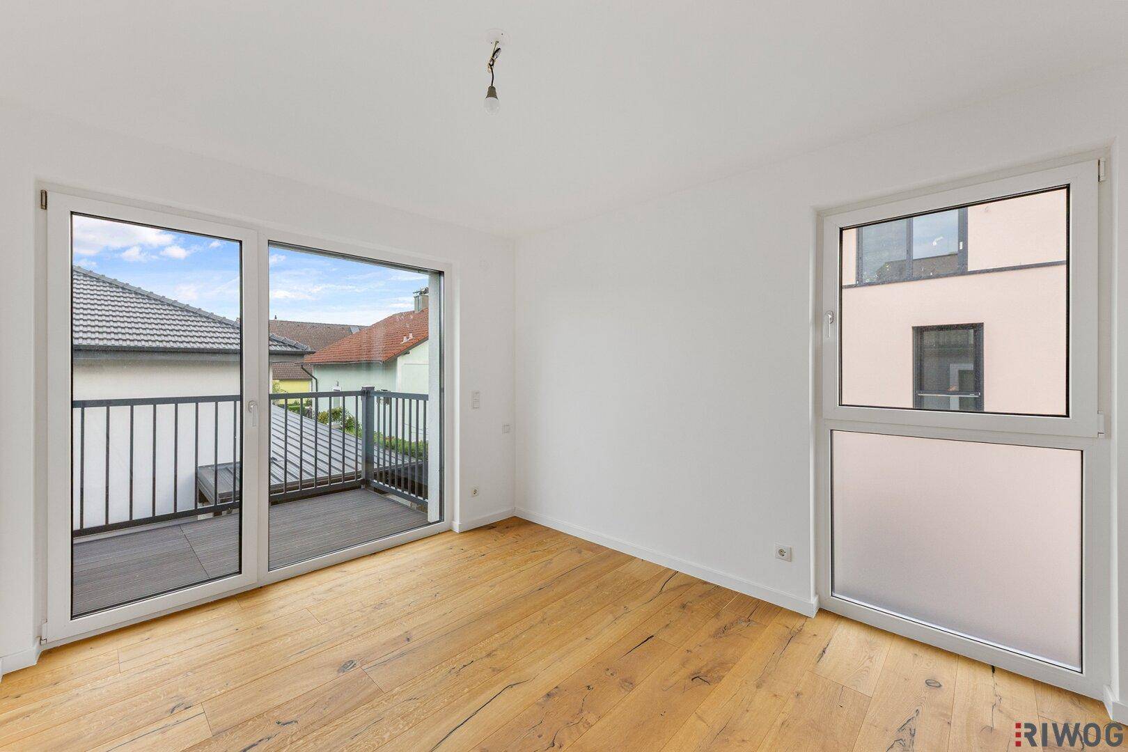 Leeres Schlafzimmer mit Holzboden und großem Fenster, das zum Balkon führt.
