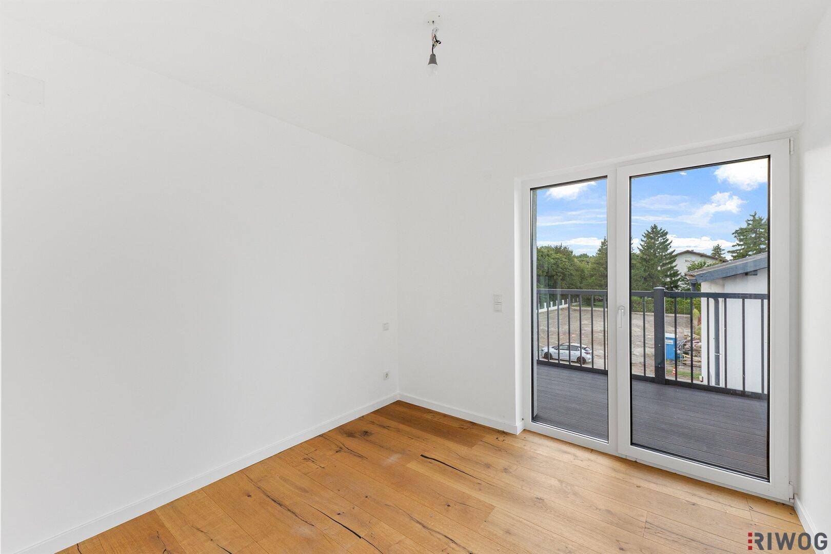 Leeres Zimmer mit Holzboden und großem Fenster, das zum Balkon führt.
