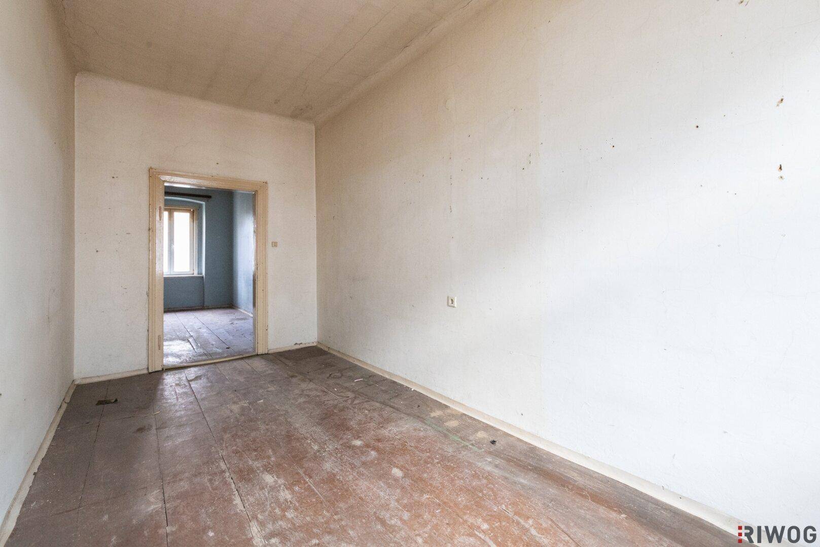 Leerer Raum mit Holzdielenboden und Durchgang zu einem weiteren Zimmer, renovierungsbedürftig.