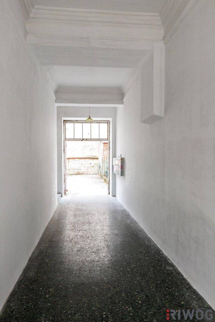Langer, schmaler Flur mit dunklem Boden und weiß gestrichenen Wänden, der zu einem Außenbereich führt.