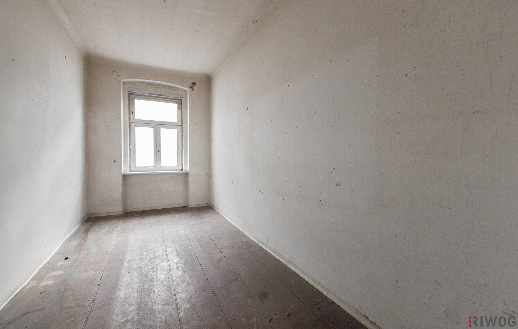 Schmales, leeres Zimmer mit Holzdielen und einem Fenster, das Renovierungsbedarf aufweist.
