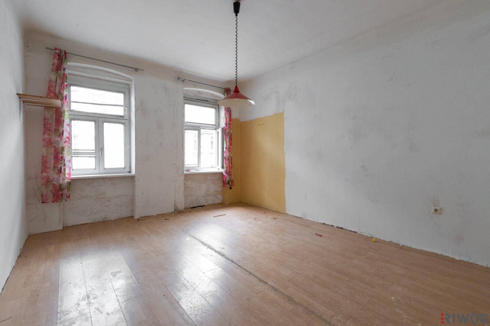 Leeres Zimmer mit alten Holzdielen, zwei Fenstern und Spuren von Renovierungsbedarf.