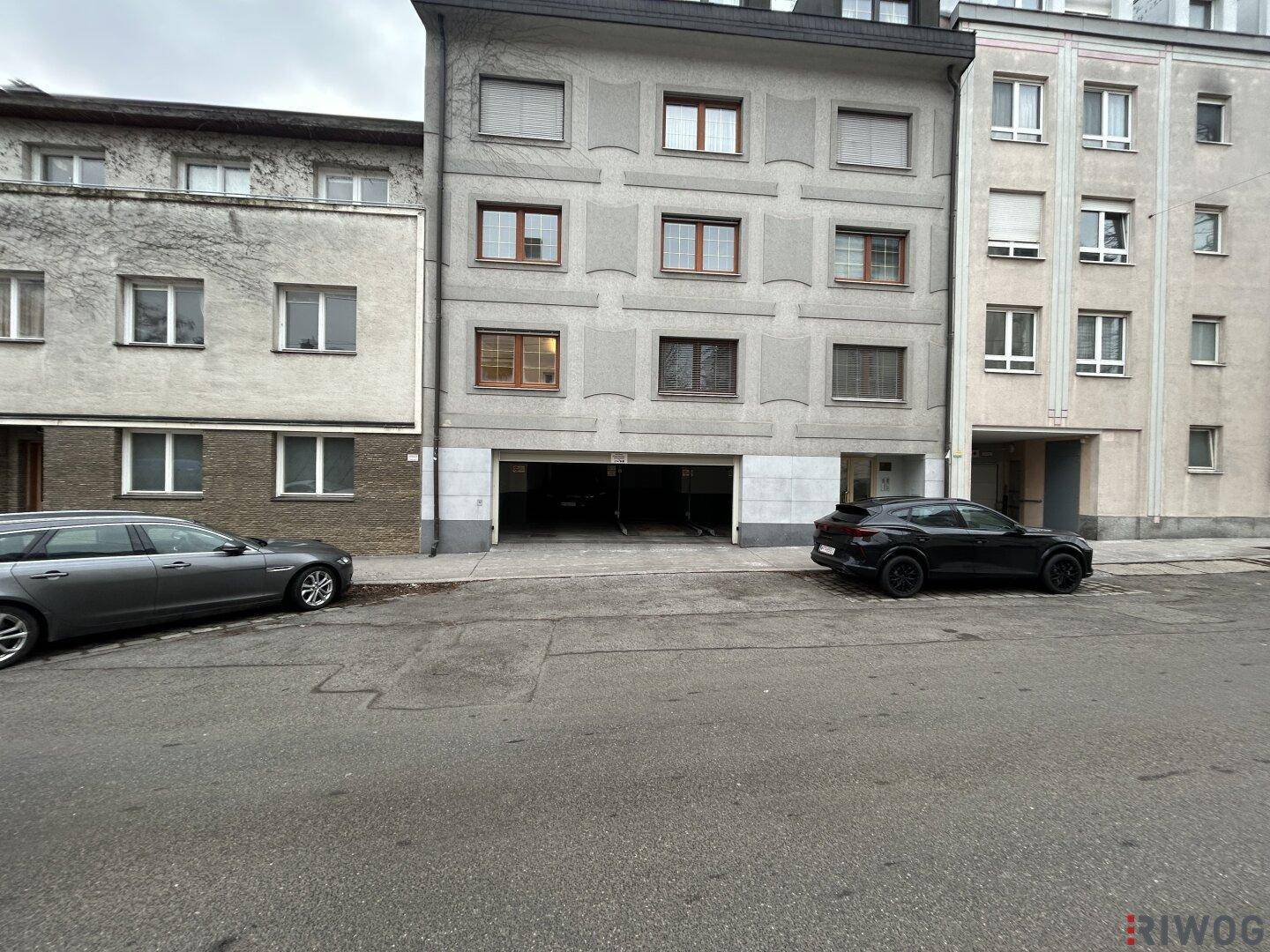 Weitwinkelansicht der Gebäudefassade mit offener Garageneinfahrt und geparkten Autos auf der Straße.