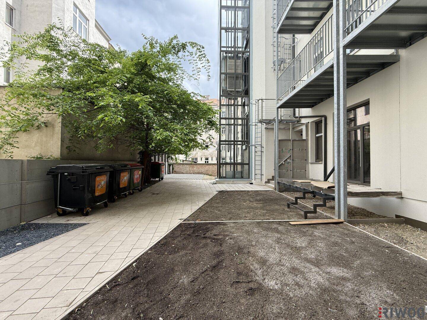Außenansicht eines Gebäudes mit externem Aufzug, gepflastertem Hof und vorbereiteter Grünfläche.