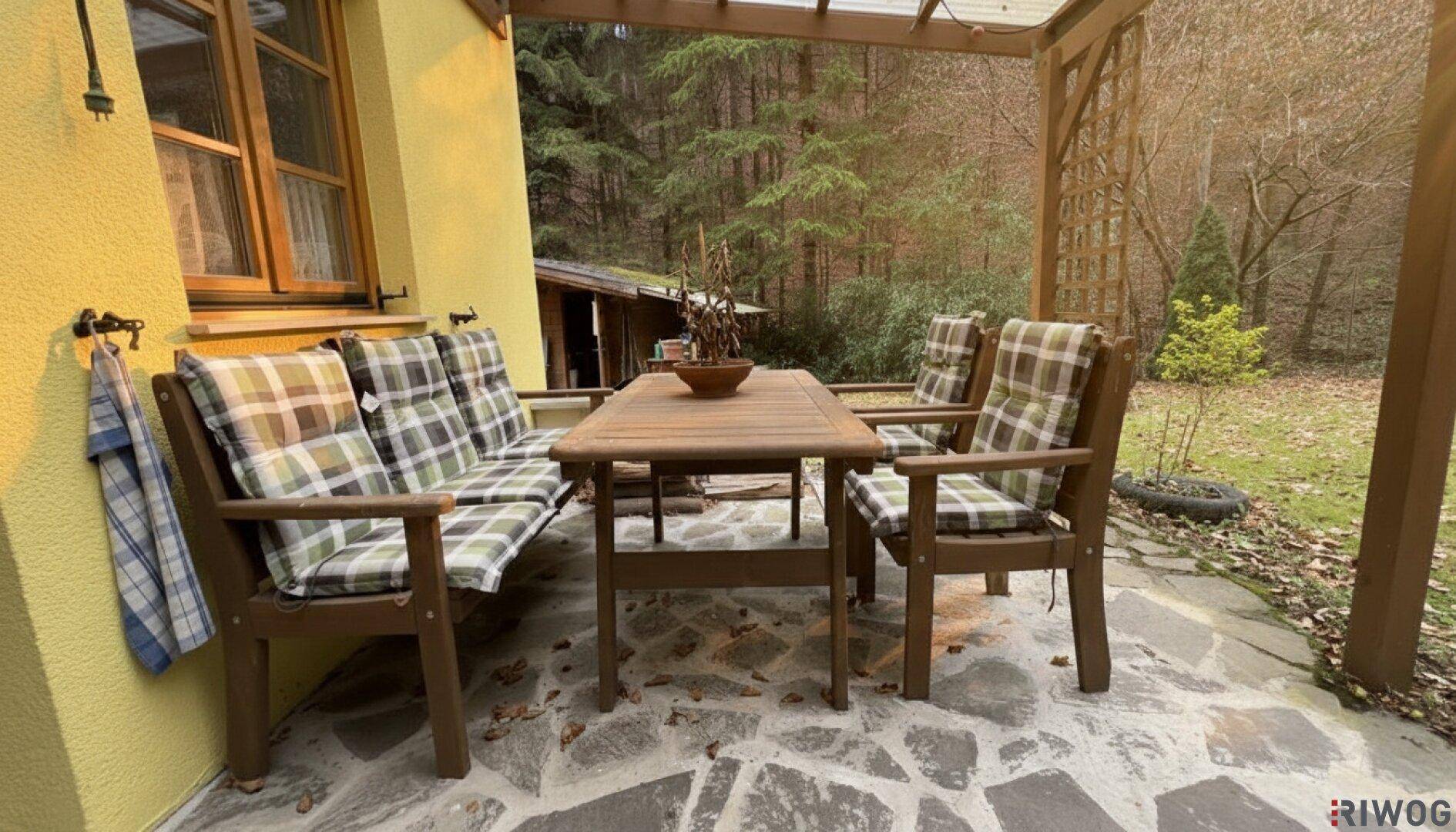 Gemütliche Terrasse mit Holzmöbeln und karierten Kissen, umgeben von Wald und Natur.