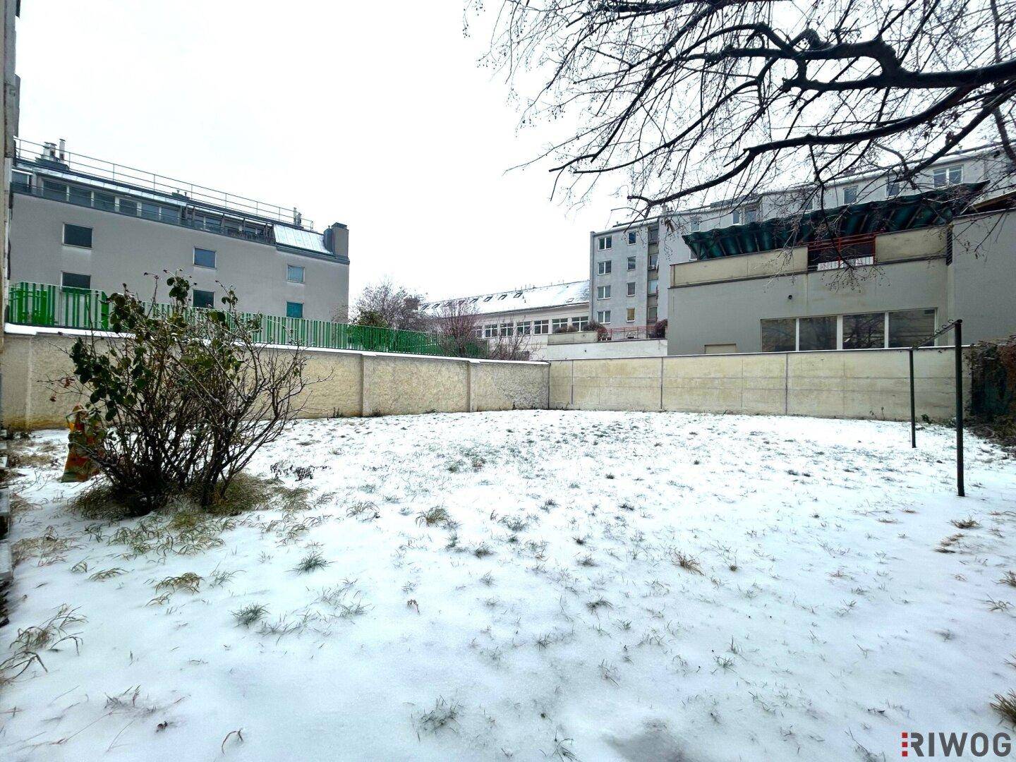 Großer Gartenbereich mit Schnee bedeckt, umgeben von einer Mauer und Blick auf umliegende Gebäude.