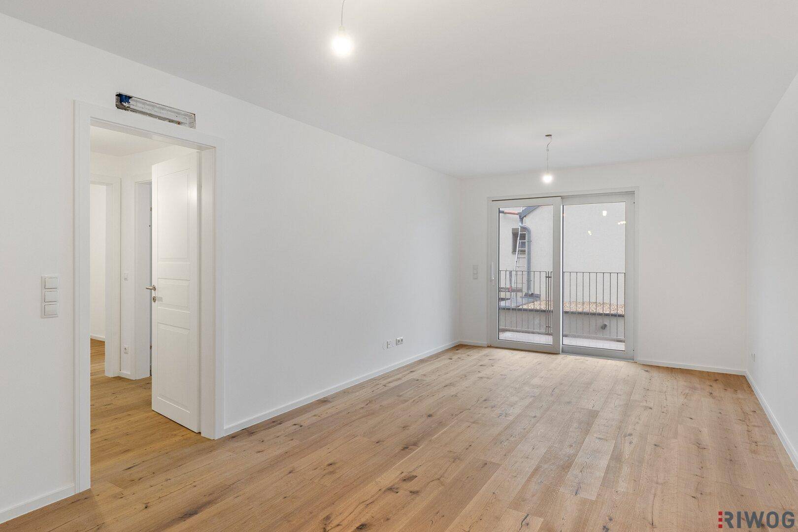 Leeres Wohnzimmer mit hellem Holzboden und Zugang zu einem französischen Balkon.