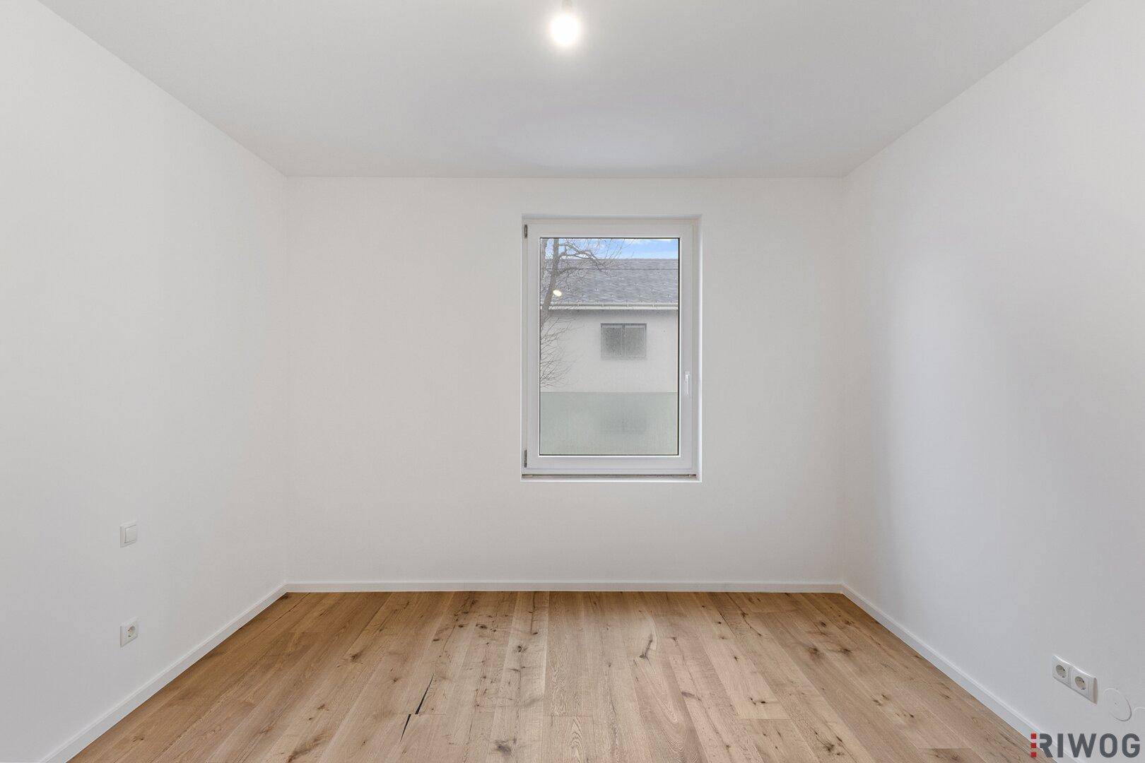 Leeres Schlafzimmer mit Holzboden und Fenster, das viel Tageslicht hereinlässt.
