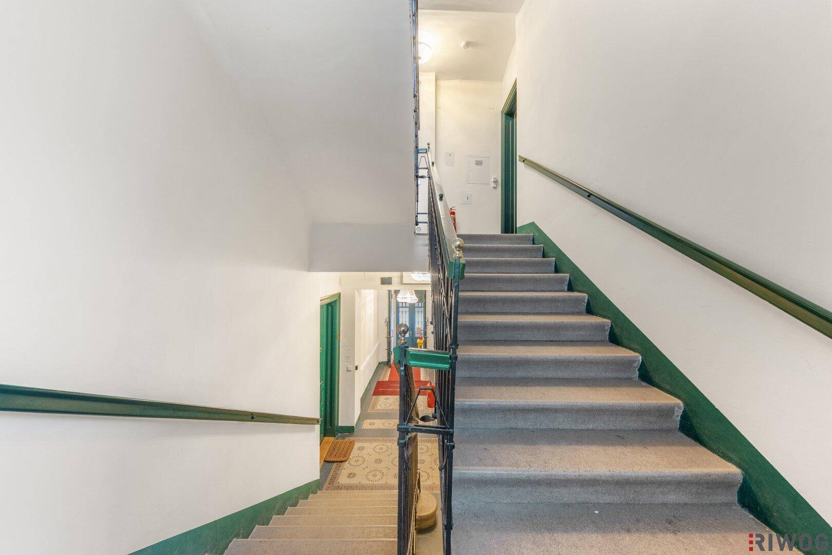Gepflegtes Treppenhaus mit Teppichstufen und grünem Handlauf in einem Mehrfamilienhaus.