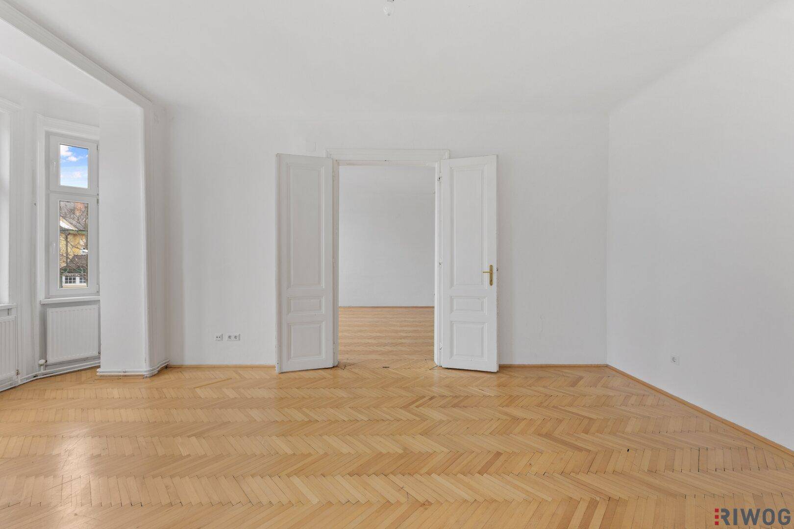 Heller Raum mit Fischgrätparkett und Flügeltüren, der zu einem weiteren Zimmer führt.