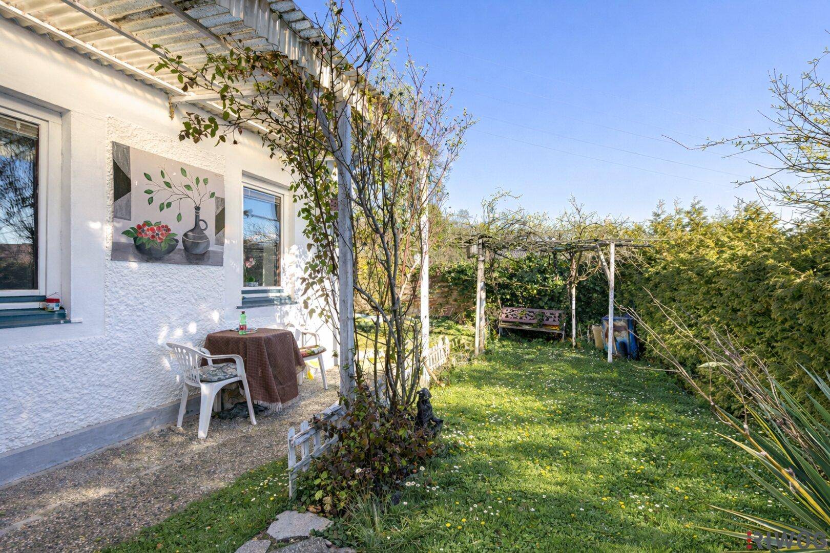 Sonniger Gartenbereich mit Schaukel und kleiner Terrasse, umgeben von Hecken und Grünflächen.