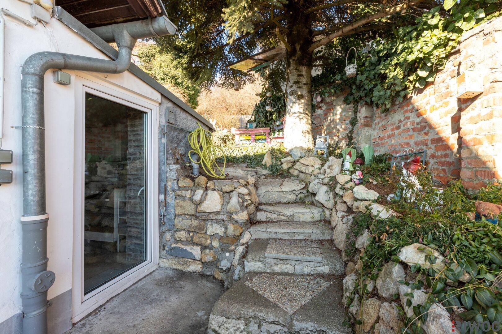 Rustikale Steintreppe führt durch den Gartenbereich, umgeben von Pflanzen und einer Ziegelmauer.