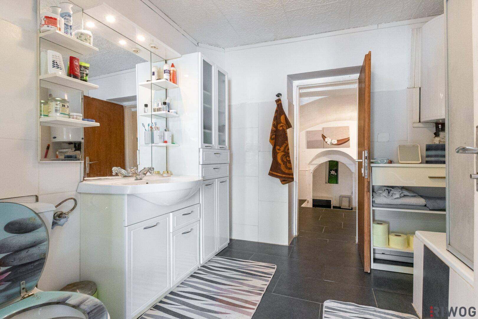 Badezimmer mit Waschtisch, Spiegelschrank und Fliesenboden, angrenzend an einen weiteren Raum.
