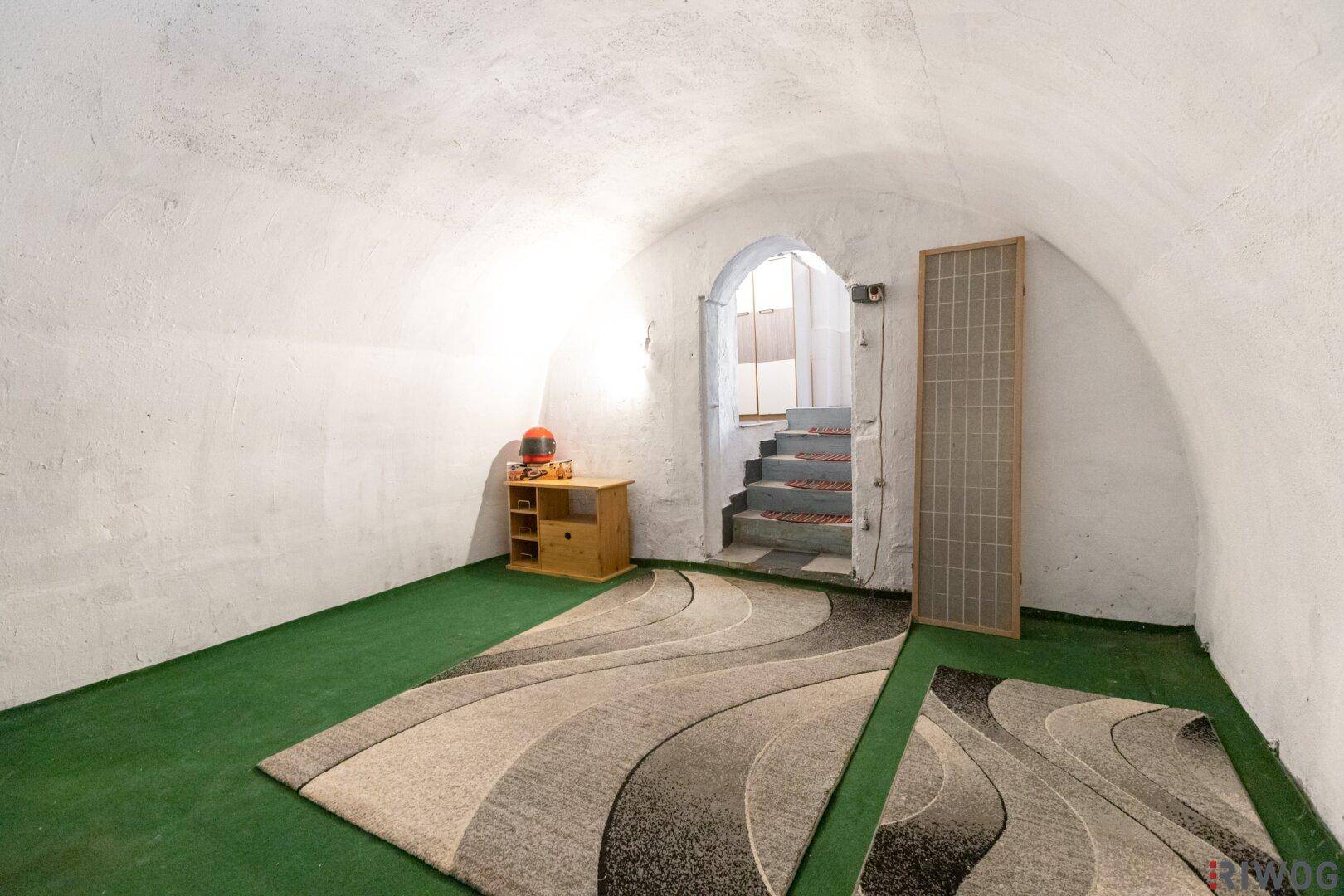 Ausgebauter Kellerraum mit grünem Teppichboden und Zugang zu einer Treppe.