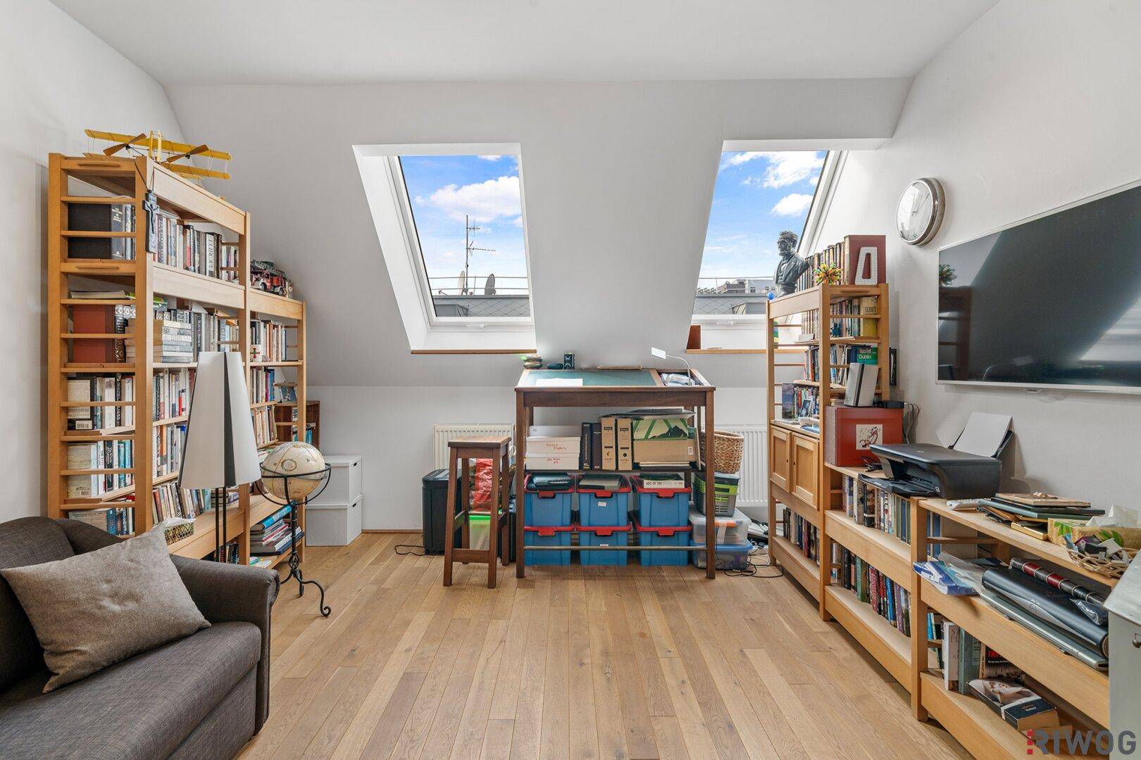 Gemütlicher Wohnbereich im Dachgeschoss mit Bücherregalen, Holzboden und zwei Dachfenstern.