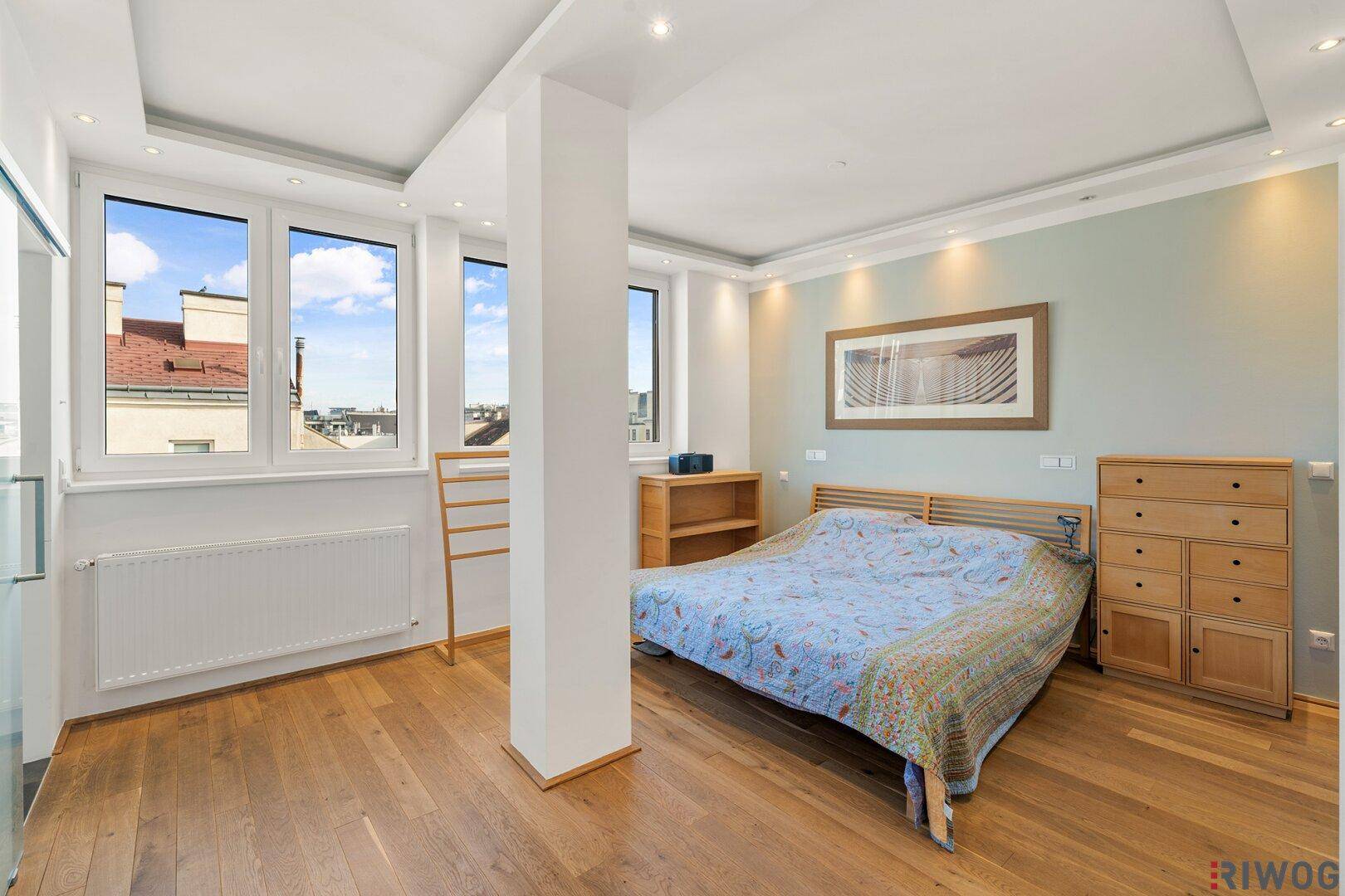 Helles Schlafzimmer mit Holzboden, großen Fenstern und Blick auf die Stadt.