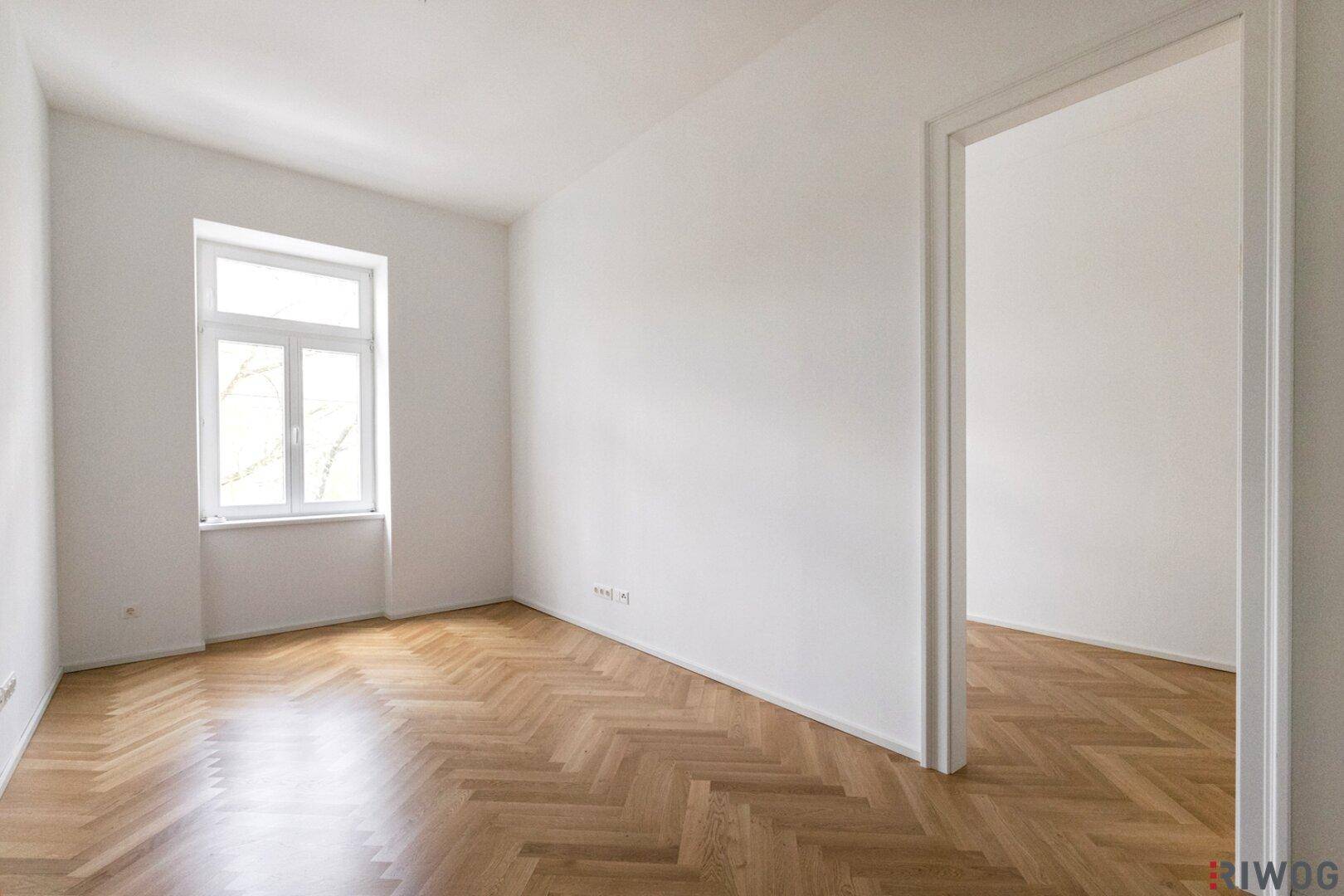 Lichtdurchfluteter Raum mit elegantem Fischgrätparkett und Blick ins Grüne durch das Fenster.