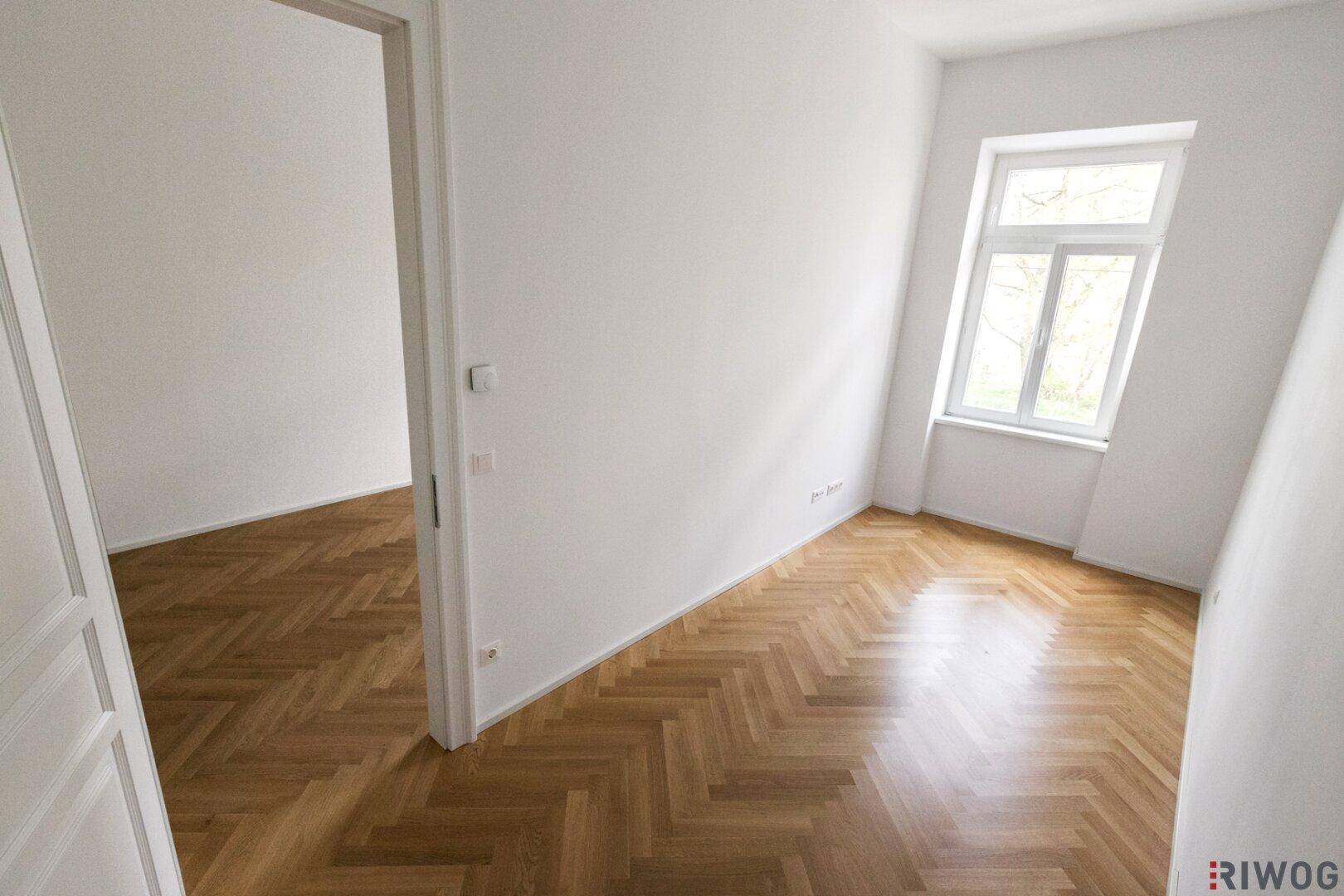 Heller Raum mit Fischgrätparkett und Zugang zu einem weiteren Zimmer, ideal für flexible Nutzung.