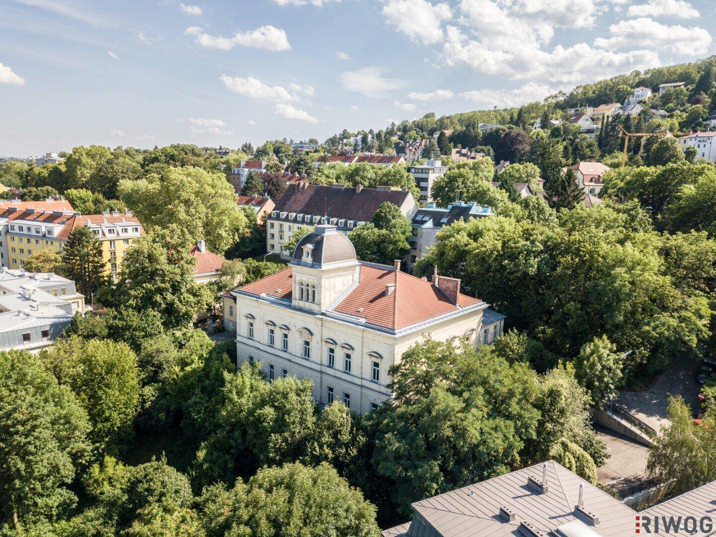 Luftaufnahme einer historischen Villa in einer grünen, hügeligen Umgebung mit vielen Bäumen.