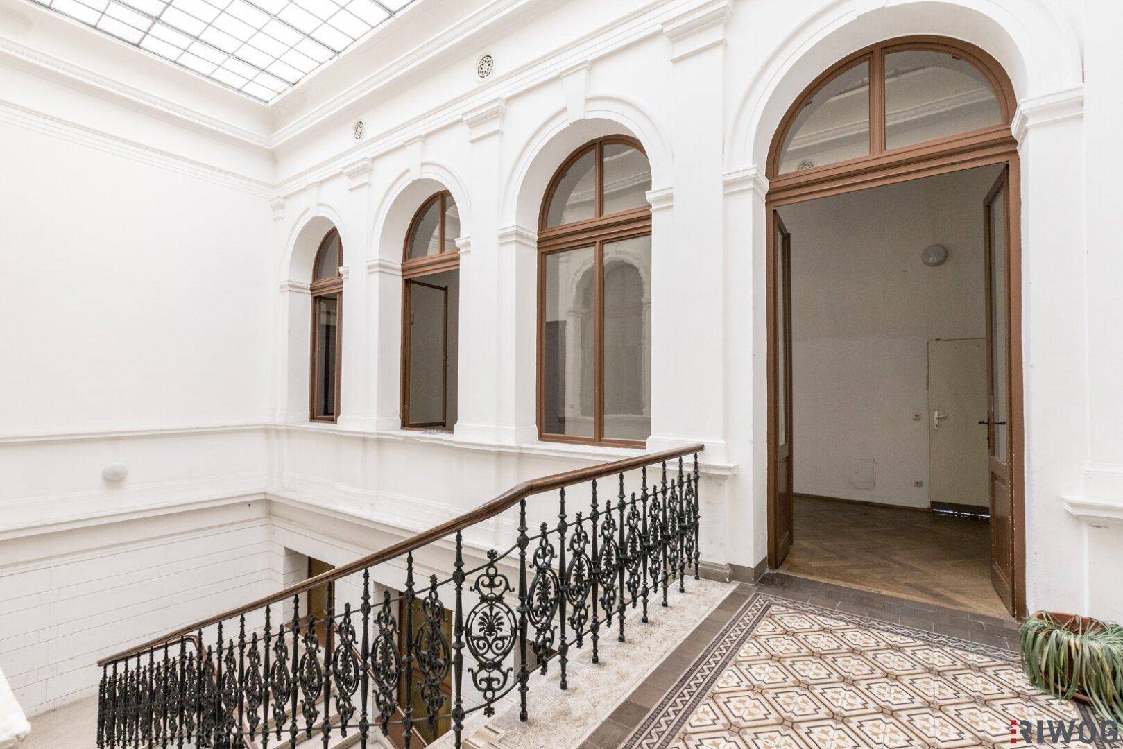 Helles Treppenhaus mit kunstvollem Geländer, hohen Bogenfenstern und Oberlicht.